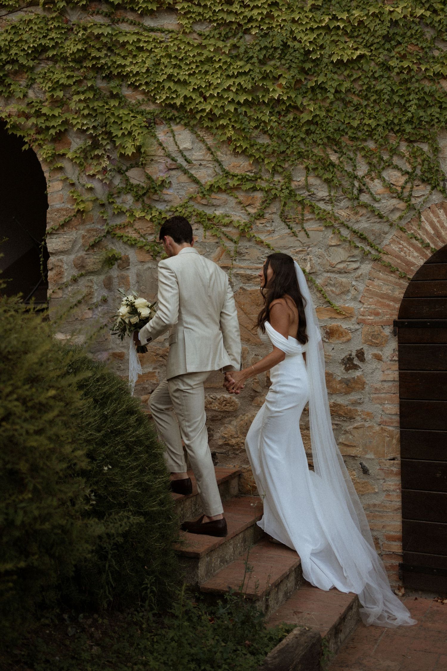 A couple in wedding attire climbs stone steps together at a rustic outdoor venue.