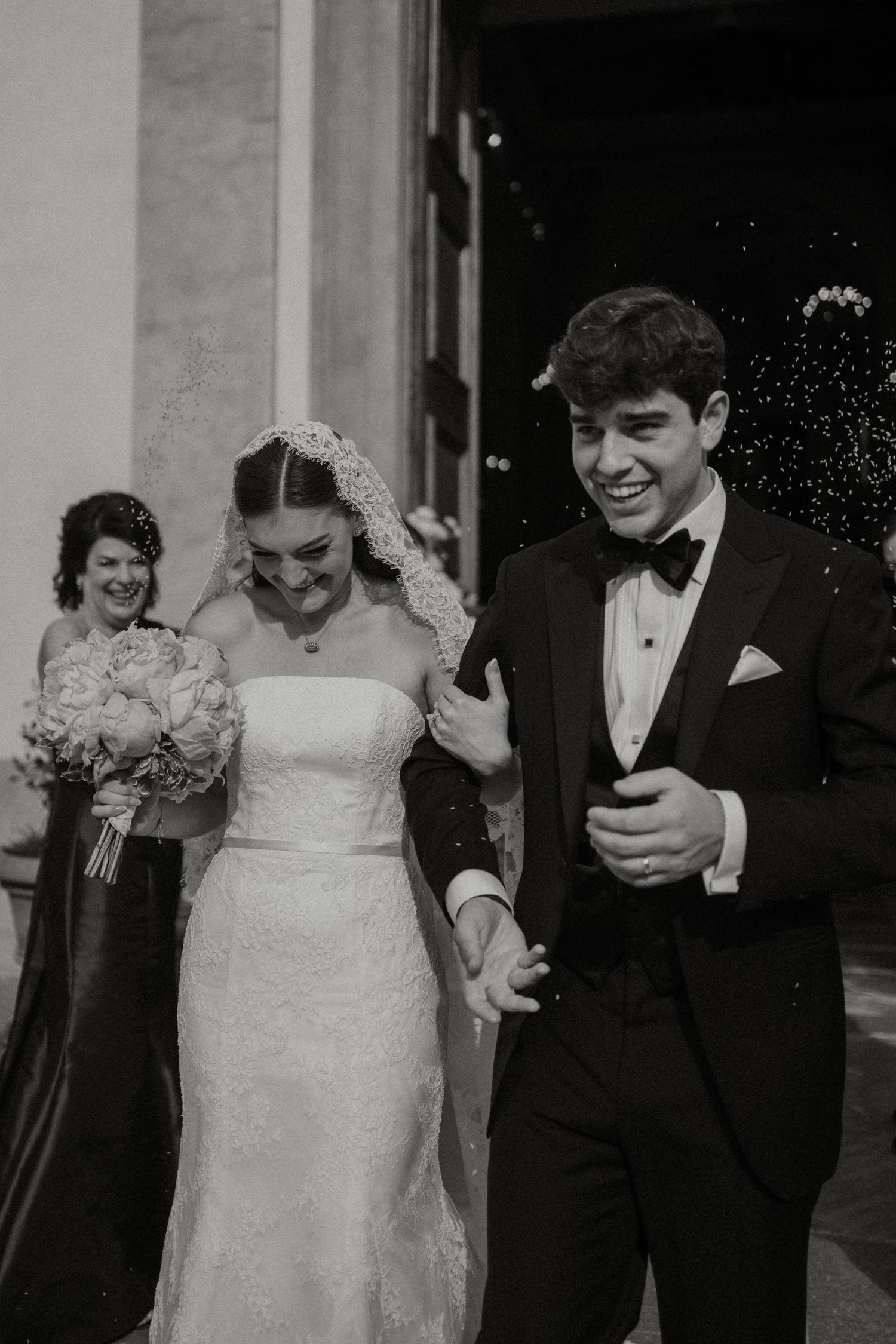Black and white photo of a smiling bride and groom exiting a church, holding hands, with guests celebrating behind them.
