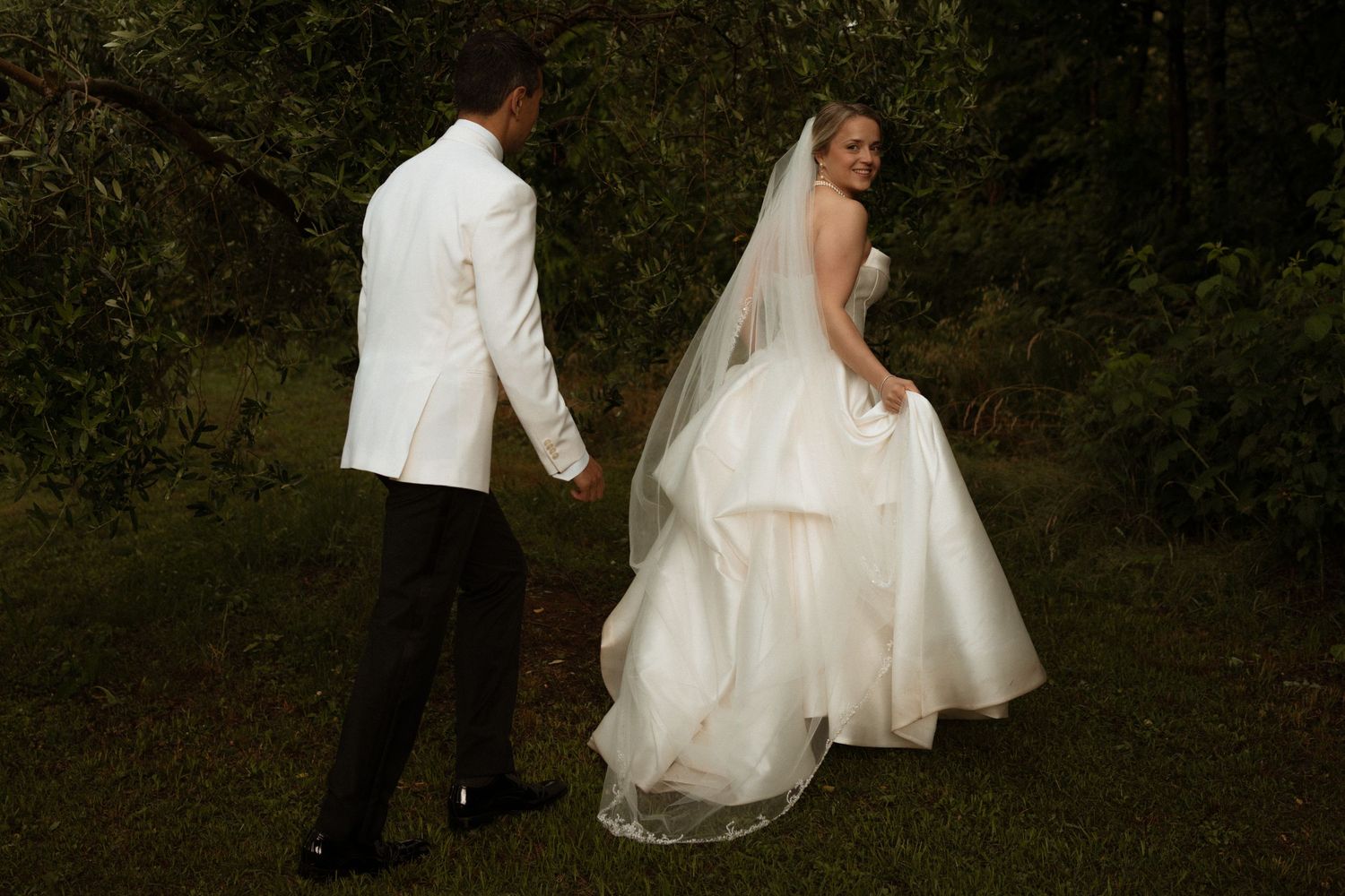 Bride in white gown and veil walks with groom in white tuxedo jacket through lush green garden setting.