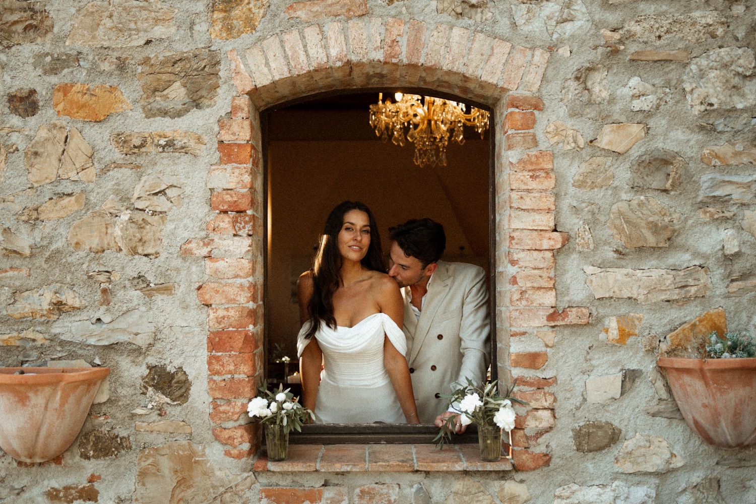 Romantic couple embraces in front of rustic stone wall with terracotta pots at Italian villa.