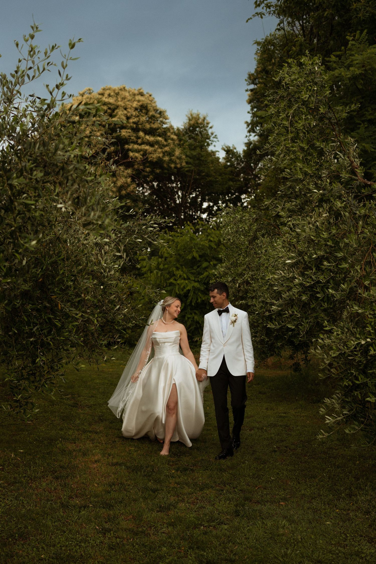 Bride and groom walk hand in hand through lush garden path, she in a white gown with veil, he in a white tuxedo jacket.
