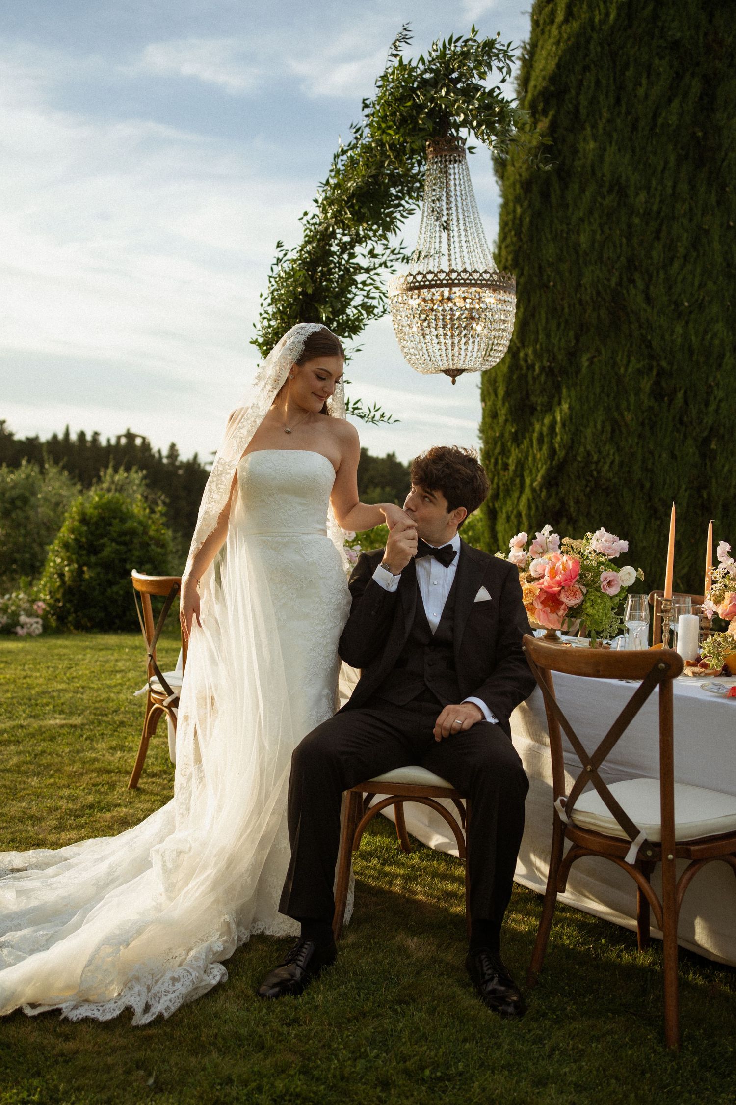 Groom in tuxedo kisses bride's hand at outdoor garden wedding reception with floral arch and chandelier.