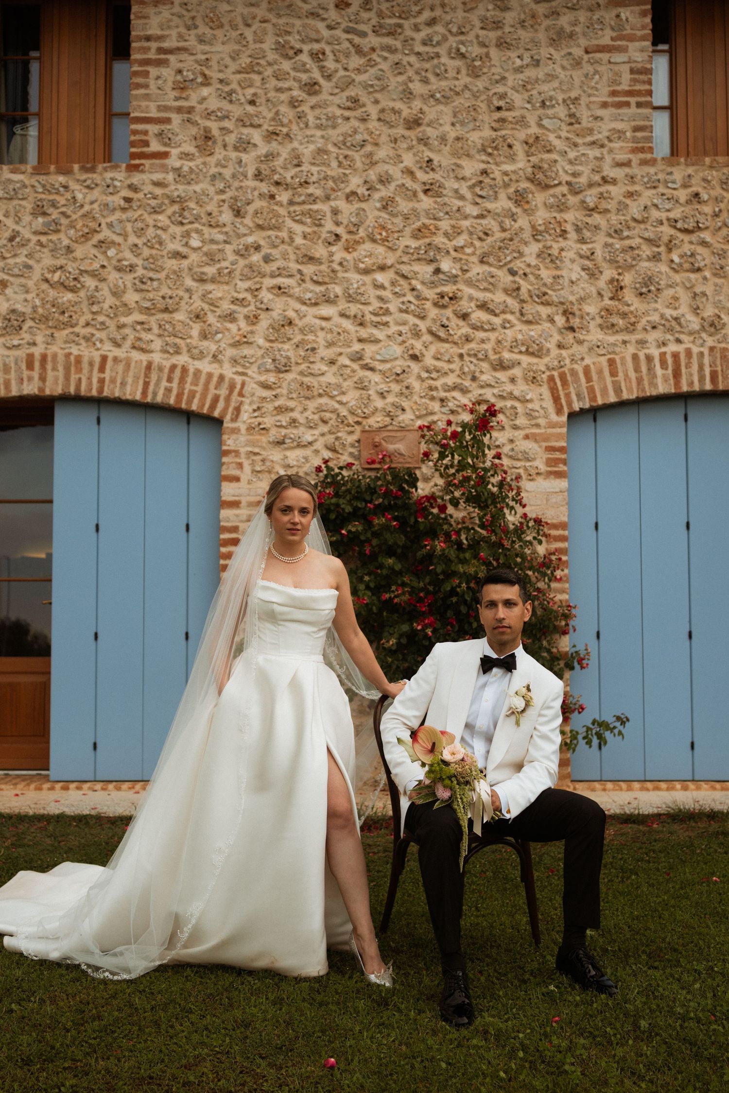 Bride in white gown and groom in white tuxedo pose before a stone building with blue shutters and red roses.