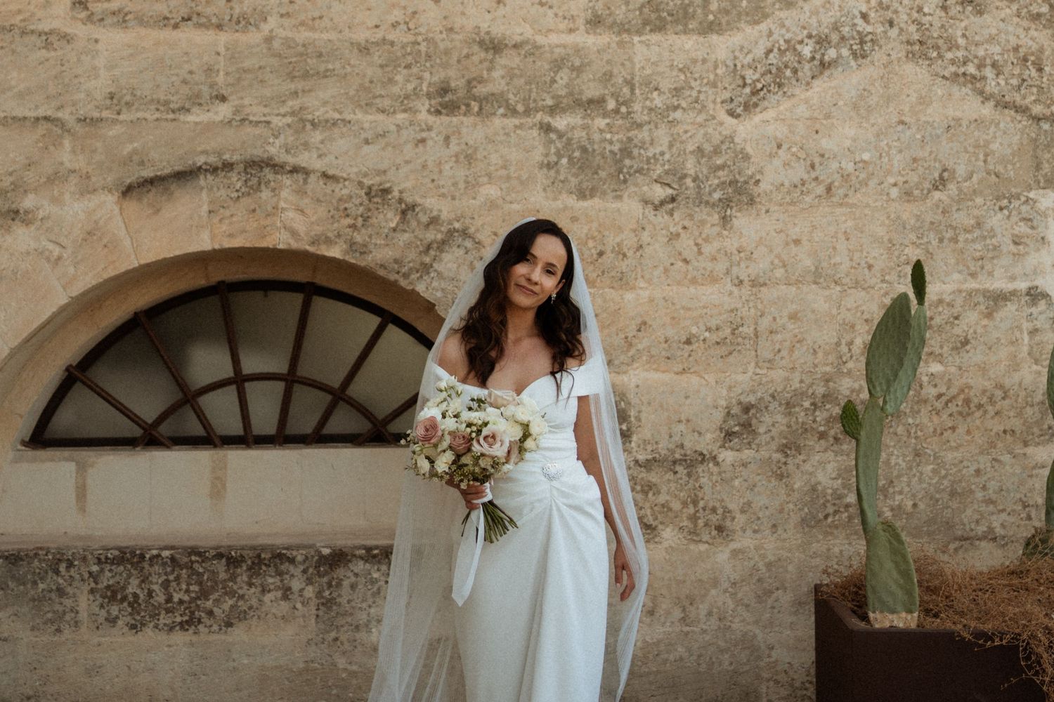 A rustic stone wall backdrop frames someone in a flowing white dress holding a bouquet of dried flowers.