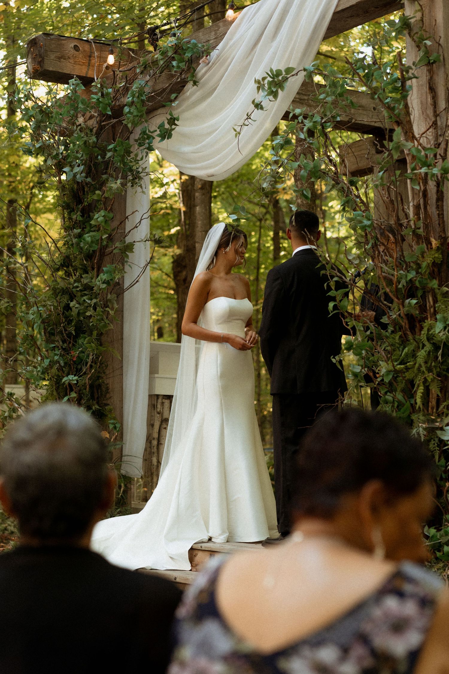 A romantic outdoor wedding ceremony takes place under a wooden arbor draped with white fabric and green vines in a forest setting.