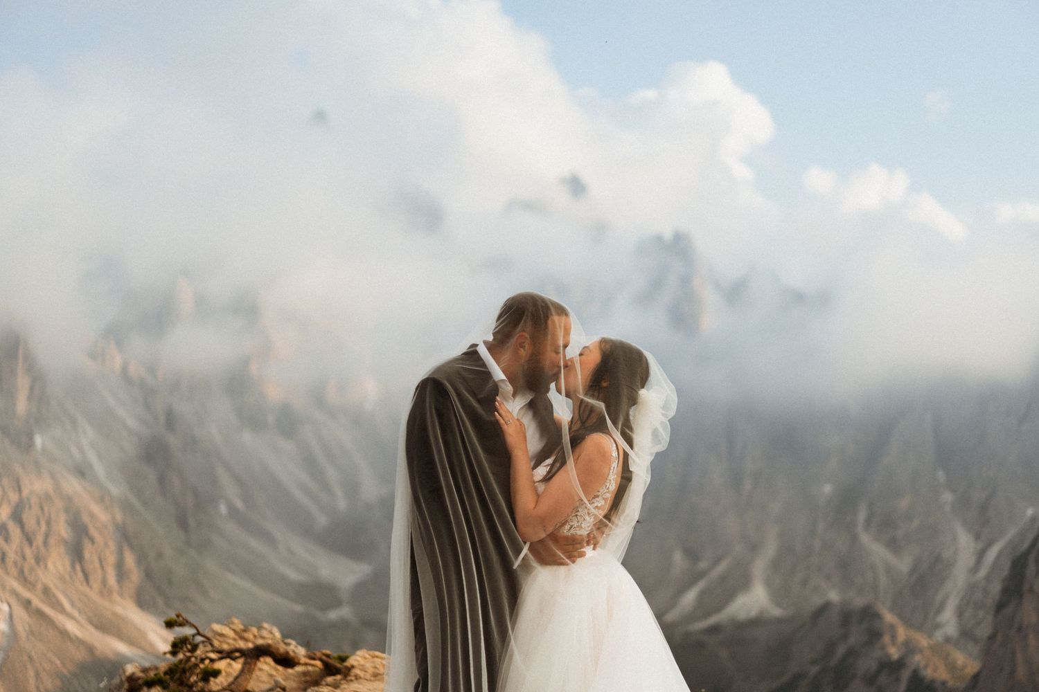 A couple in wedding attire share an intimate moment on a mountain peak with dramatic rocky peaks in the background.