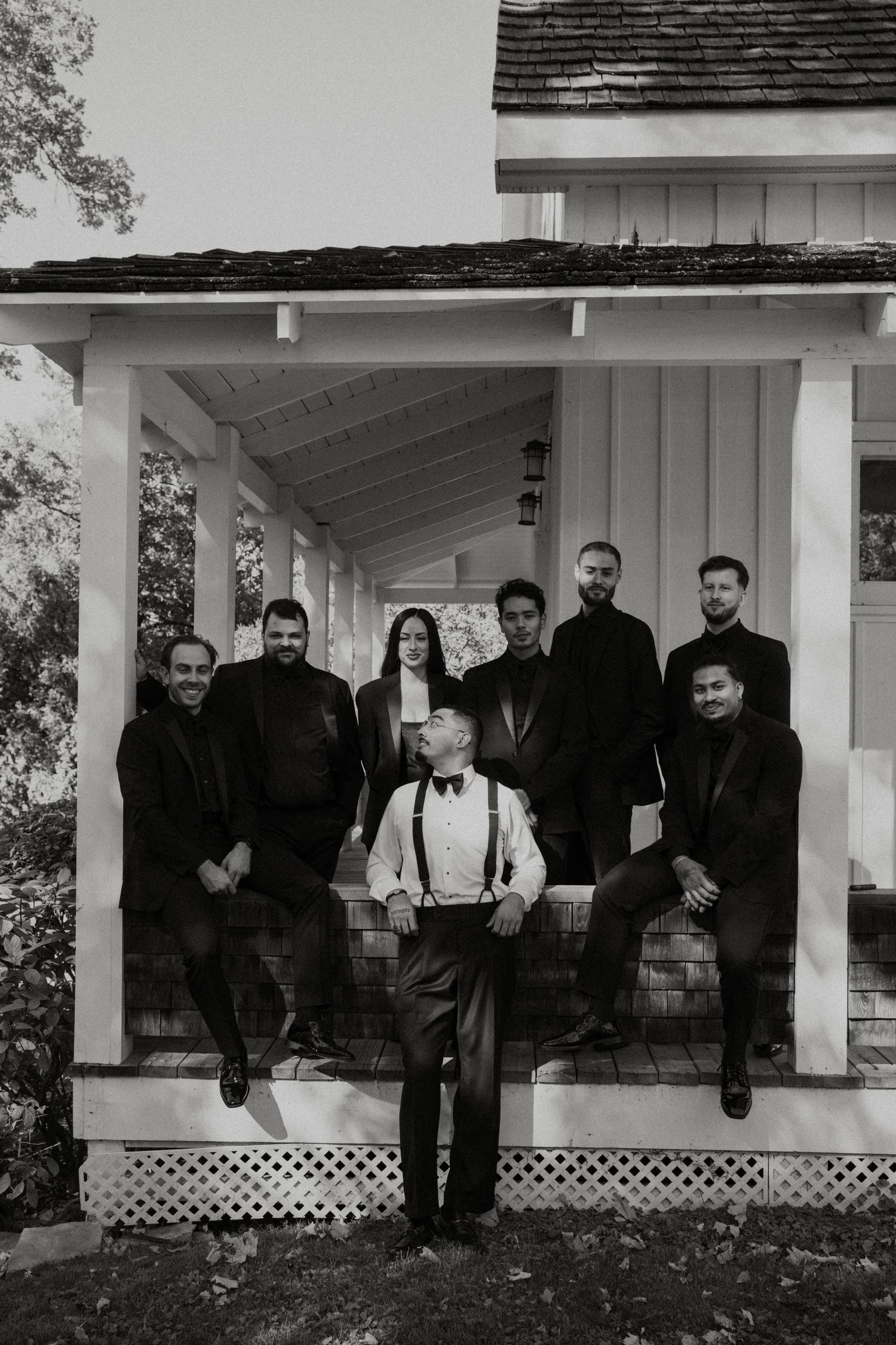 A group of formally dressed people pose together on a white porch with lattice details in black and white photography.