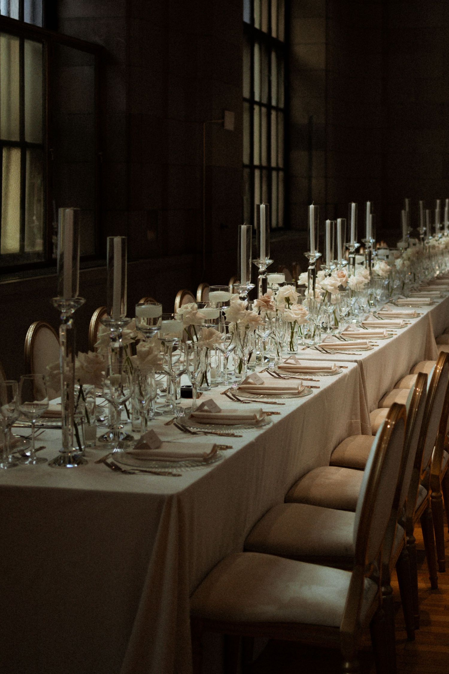 Elegant long banquet table with white linens, tall candles, floral centerpieces, and gold chairs in a dimly lit venue.