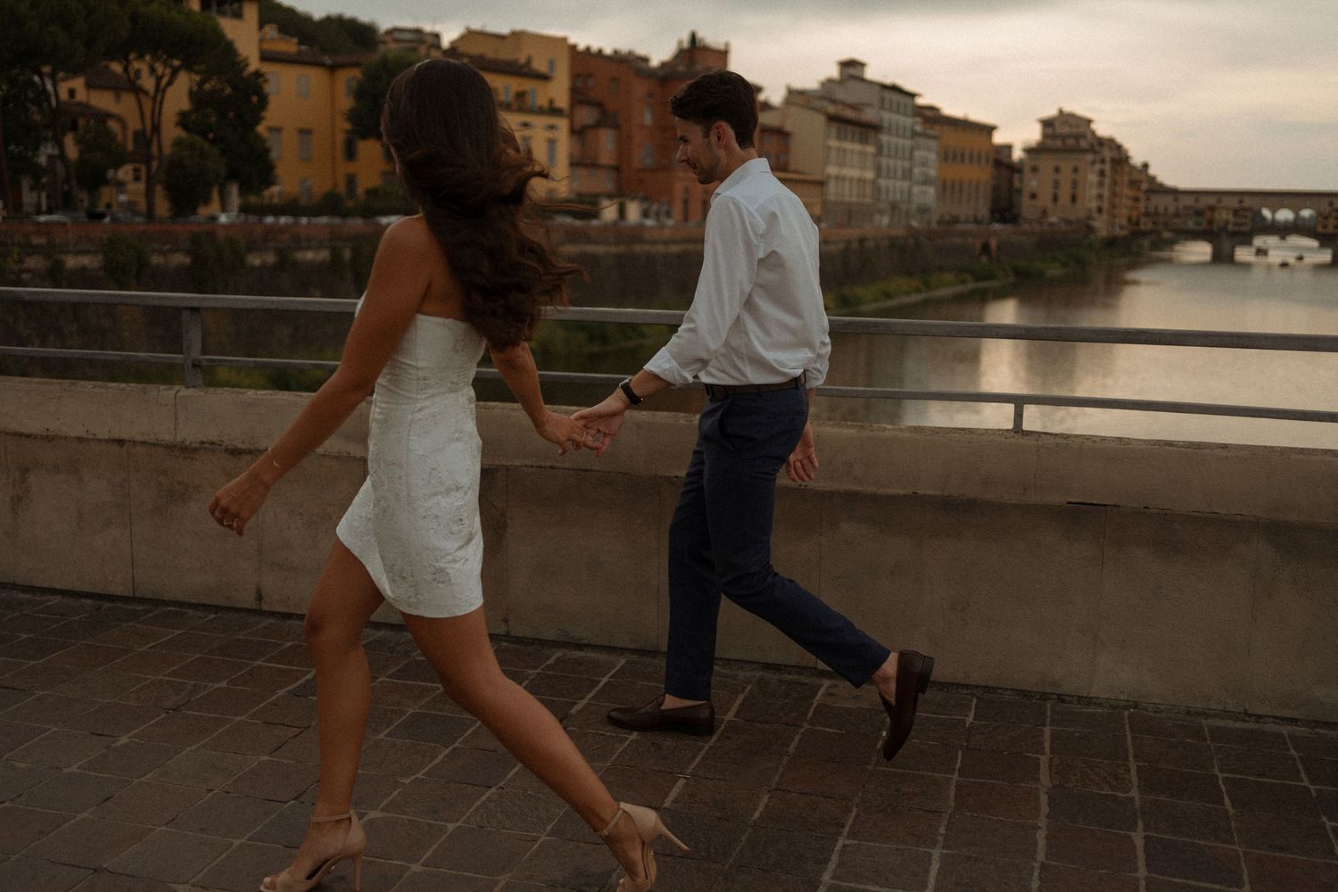 A couple walking hand in hand along a riverside promenade at sunset with historic buildings in the background.