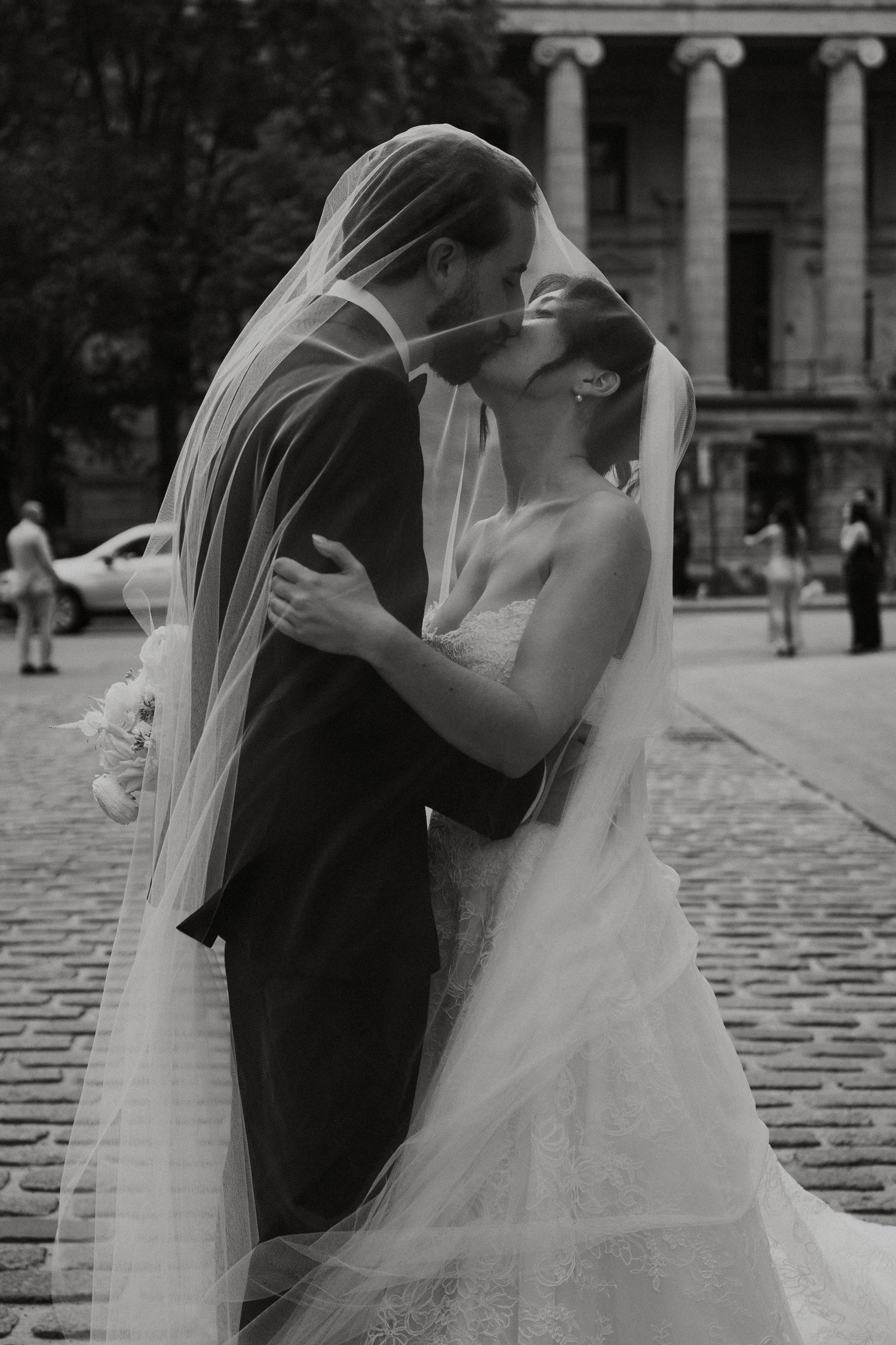 Black and white photo of bride and groom kissing under veil on cobblestone street outside historic building.
