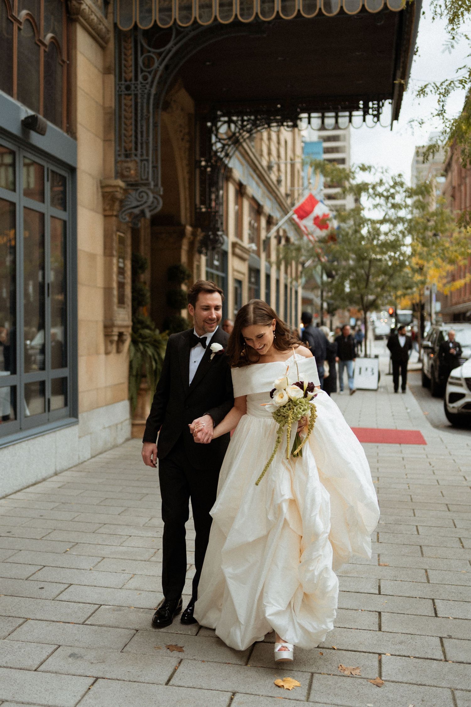 A wedding couple walks along a city sidewalk on an autumn day with fallen leaves scattered on the ground.