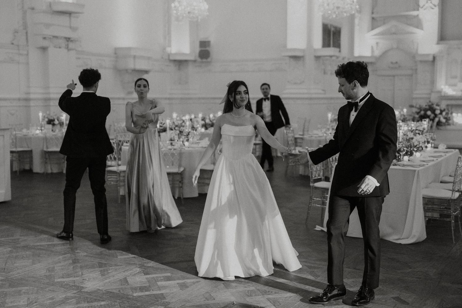 Wedding guests dancing and celebrating at an elegant reception in a white ballroom with ornate chandeliers.