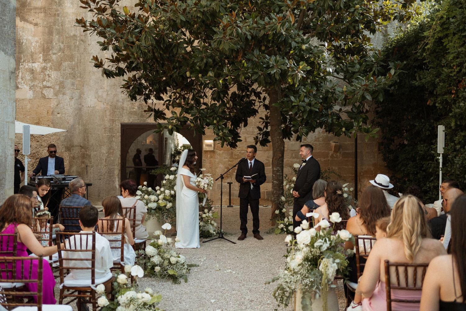 Wedding ceremony guests gather in a rustic stone courtyard decorated with elegant floral arrangements.