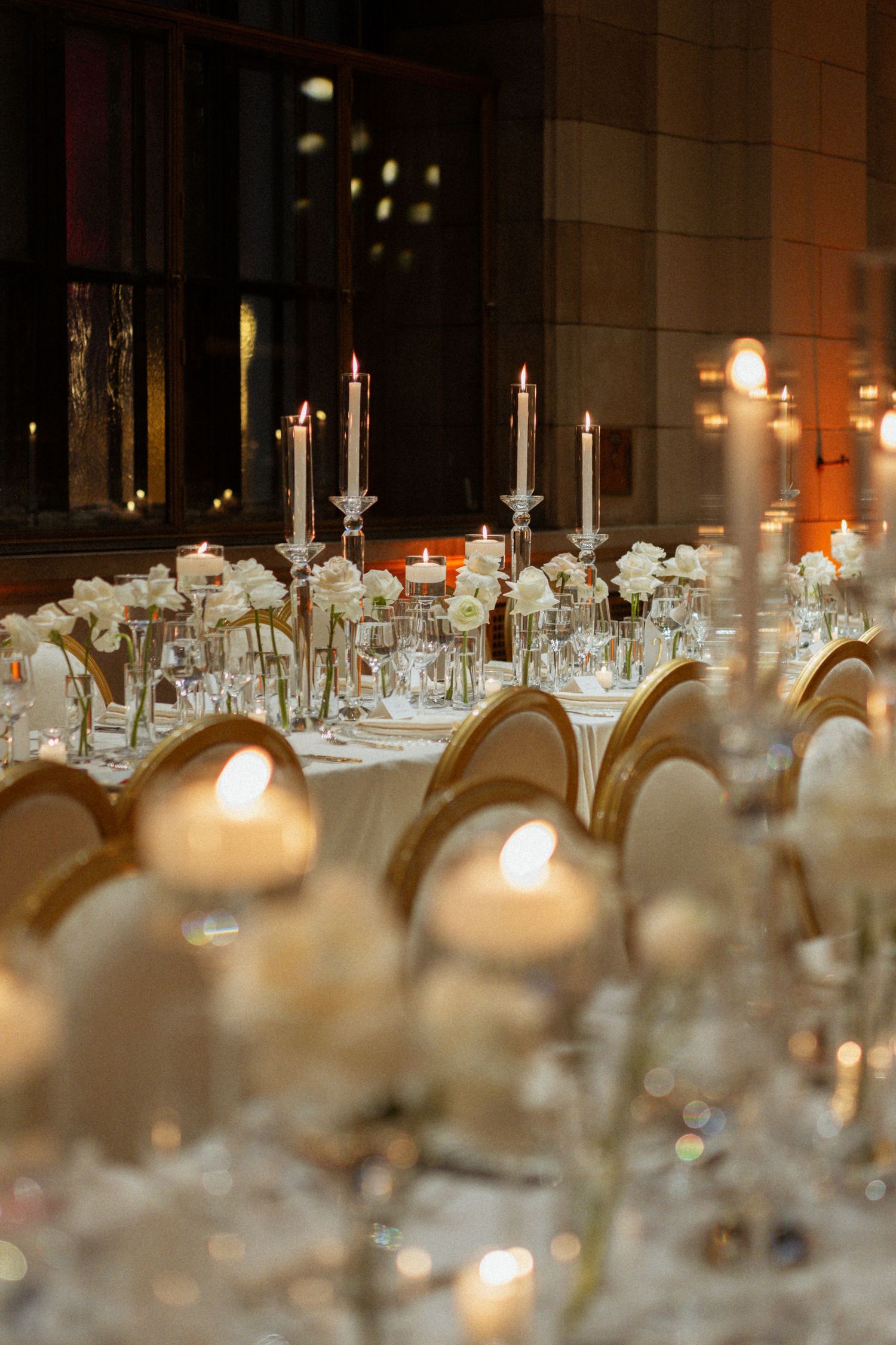 Elegant wedding reception table with crystal candelabras, white roses, glowing candles, and gold chairs in a softly lit venue.