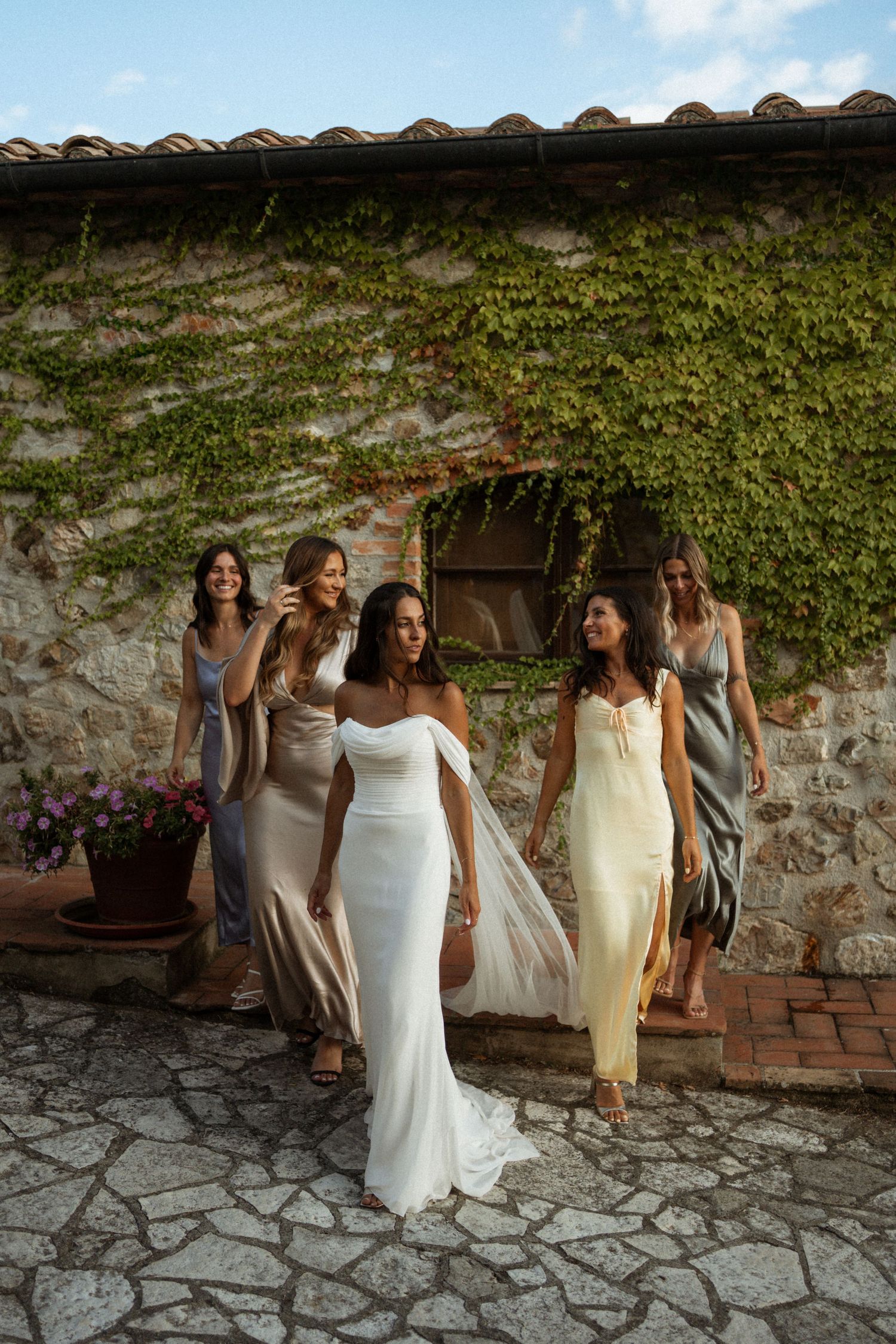 Group of bridesmaids in light-colored dresses pose together against a rustic stone wall.