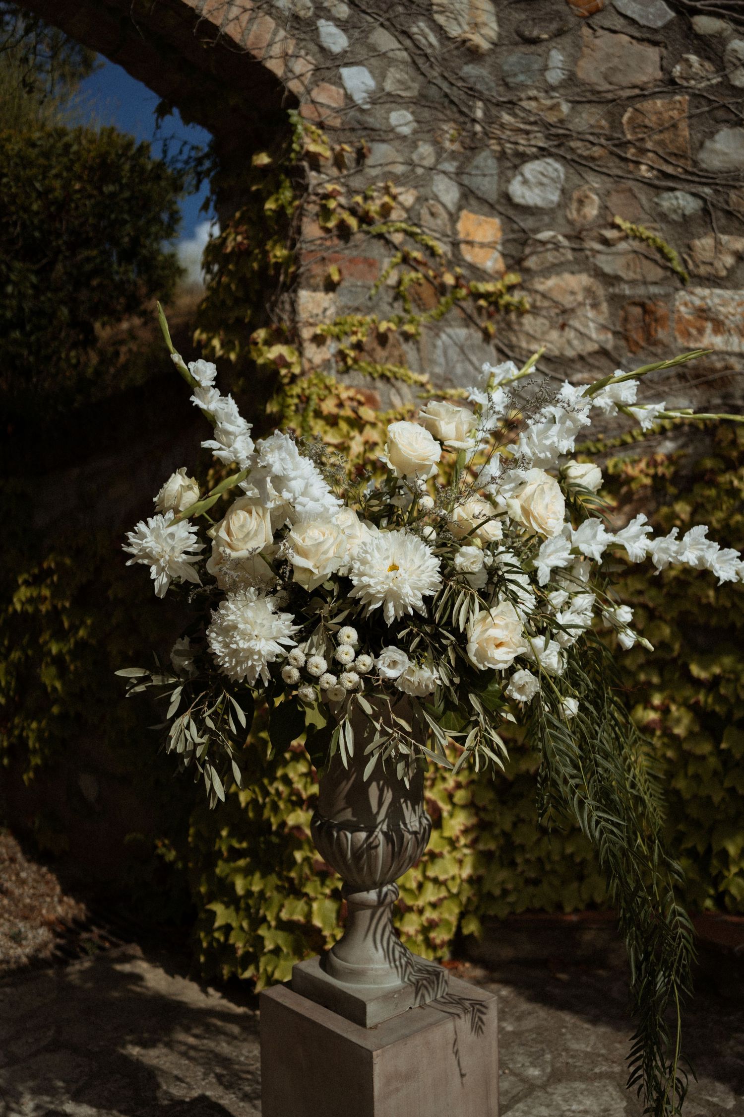 White floral arrangements in stone urns create an elegant display against a rustic stone wall in natural lighting.