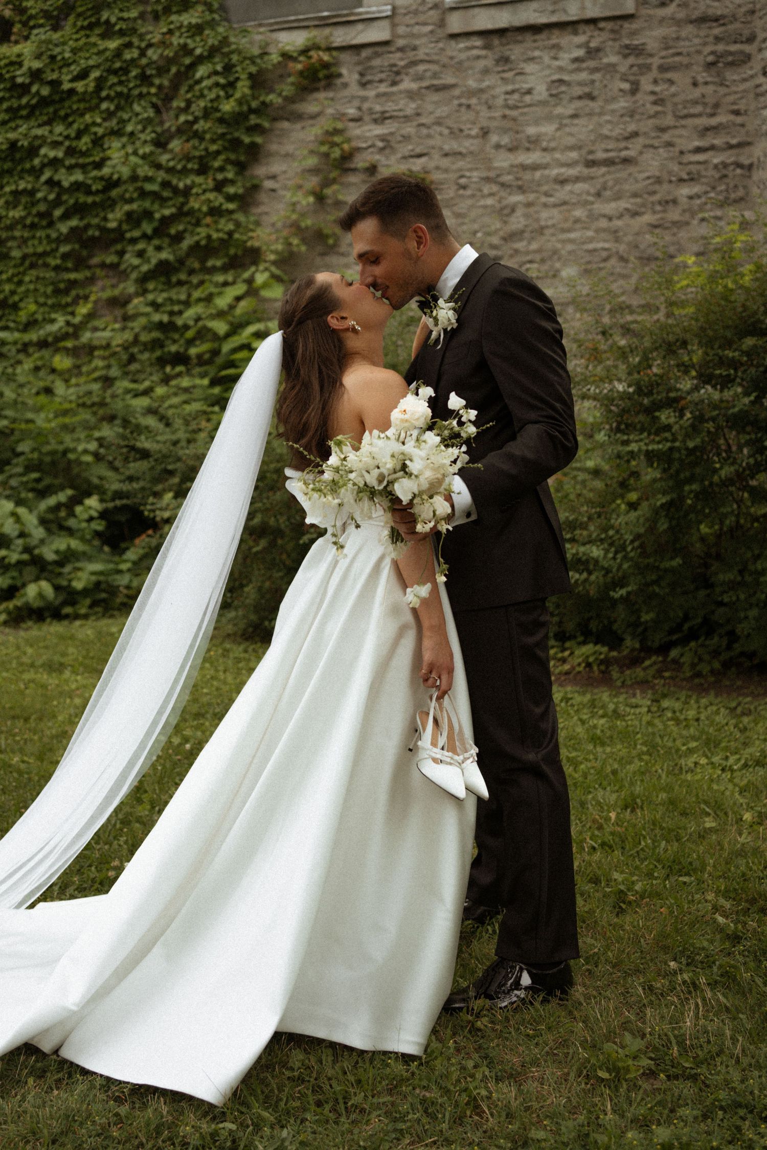 A romantic wedding couple shares a kiss against a stone wall with ivy, the bride wearing a flowing white gown with long veil.