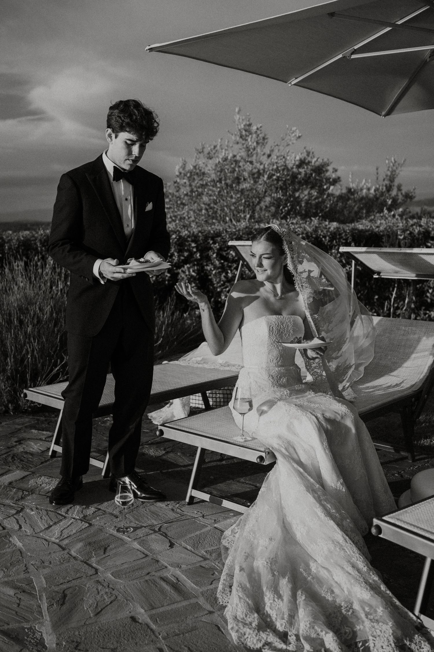 Black and white photo of a bride in lace gown and groom in tuxedo sharing a moment outdoors at their wedding.