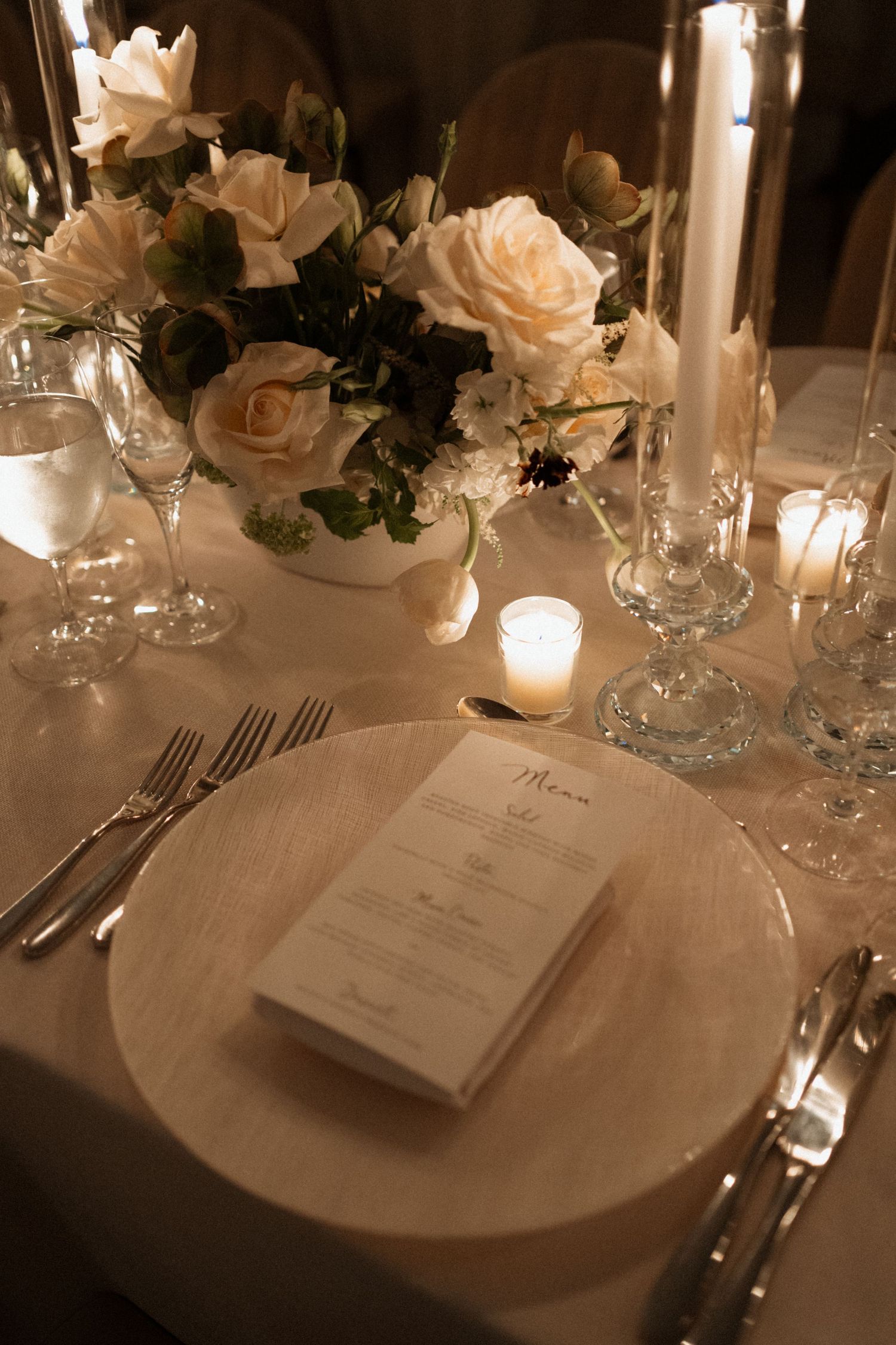 Elegant wedding reception table setting with white roses, glowing candles, crystal glassware and printed menu cards.
