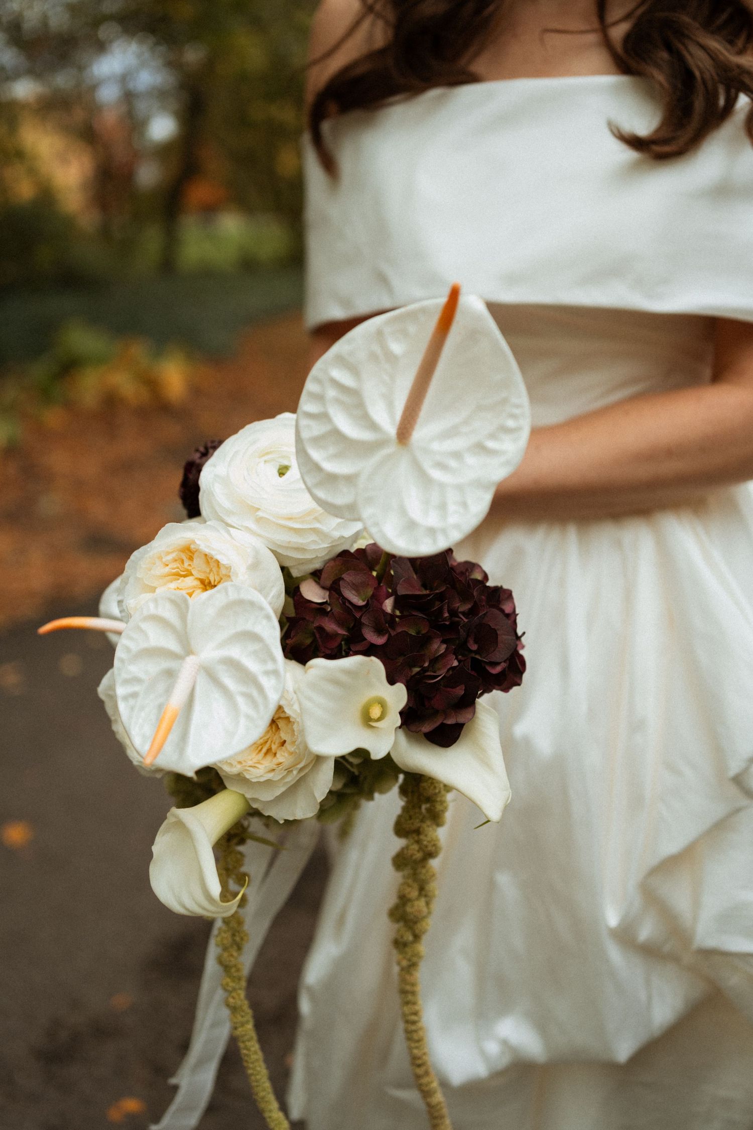 A bridal bouquet featuring white anthurium lilies, roses, and dark burgundy flowers against a white wedding dress.