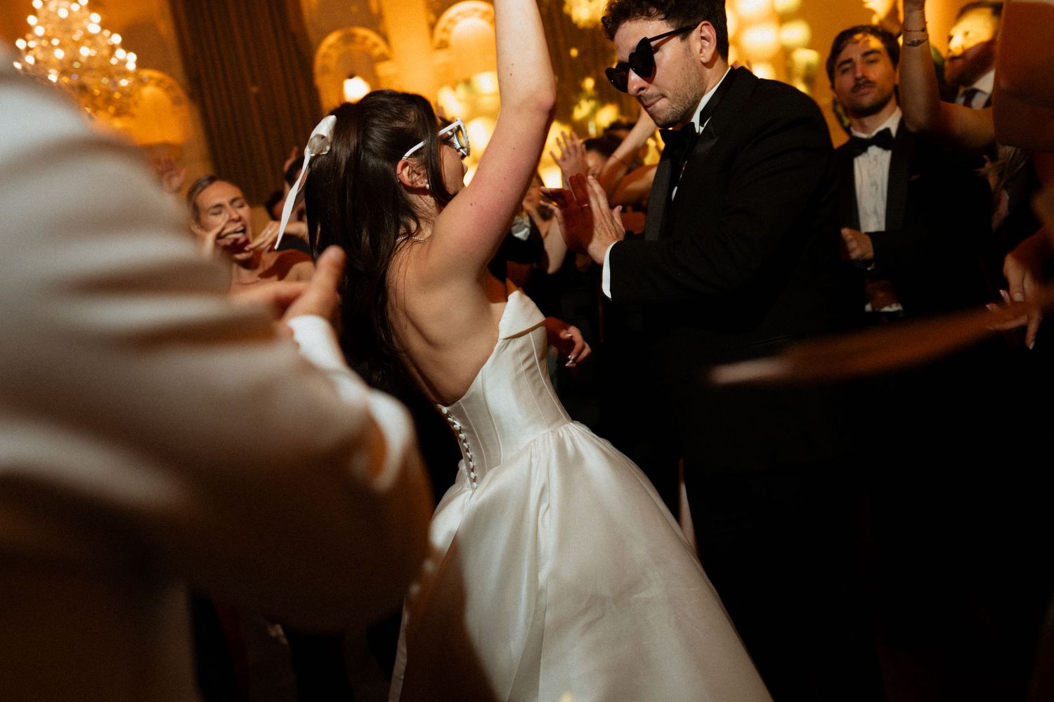 A couple shares their first dance at a wedding reception with warm lighting and guests watching in the background.