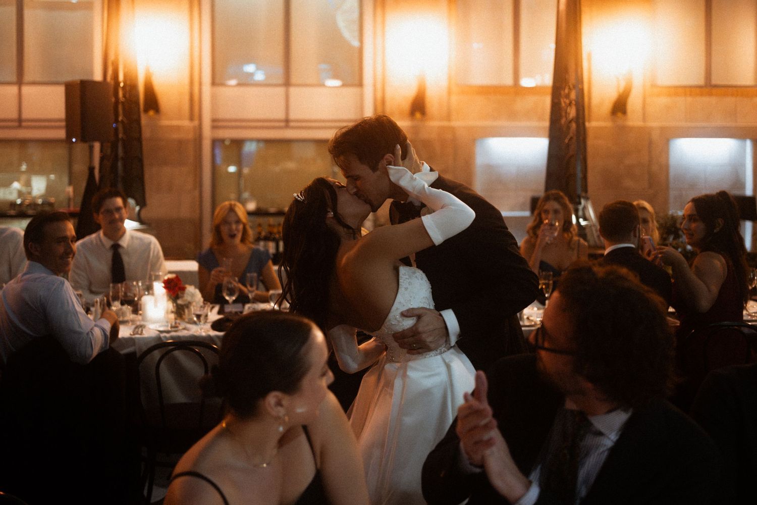 A couple shares an intimate dance moment at an elegant indoor wedding reception with warm lighting and guests seated nearby.