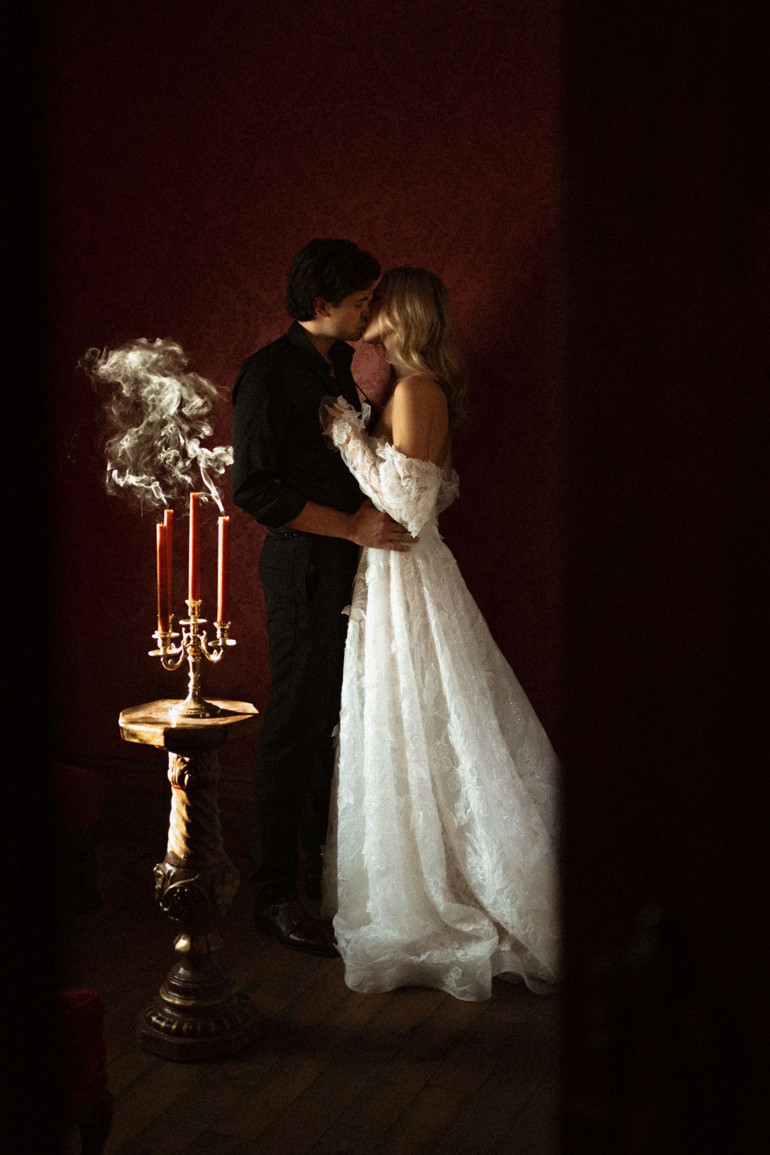 Romantic silhouette of couple in formal attire next to candlelit stand against dark red wall.
