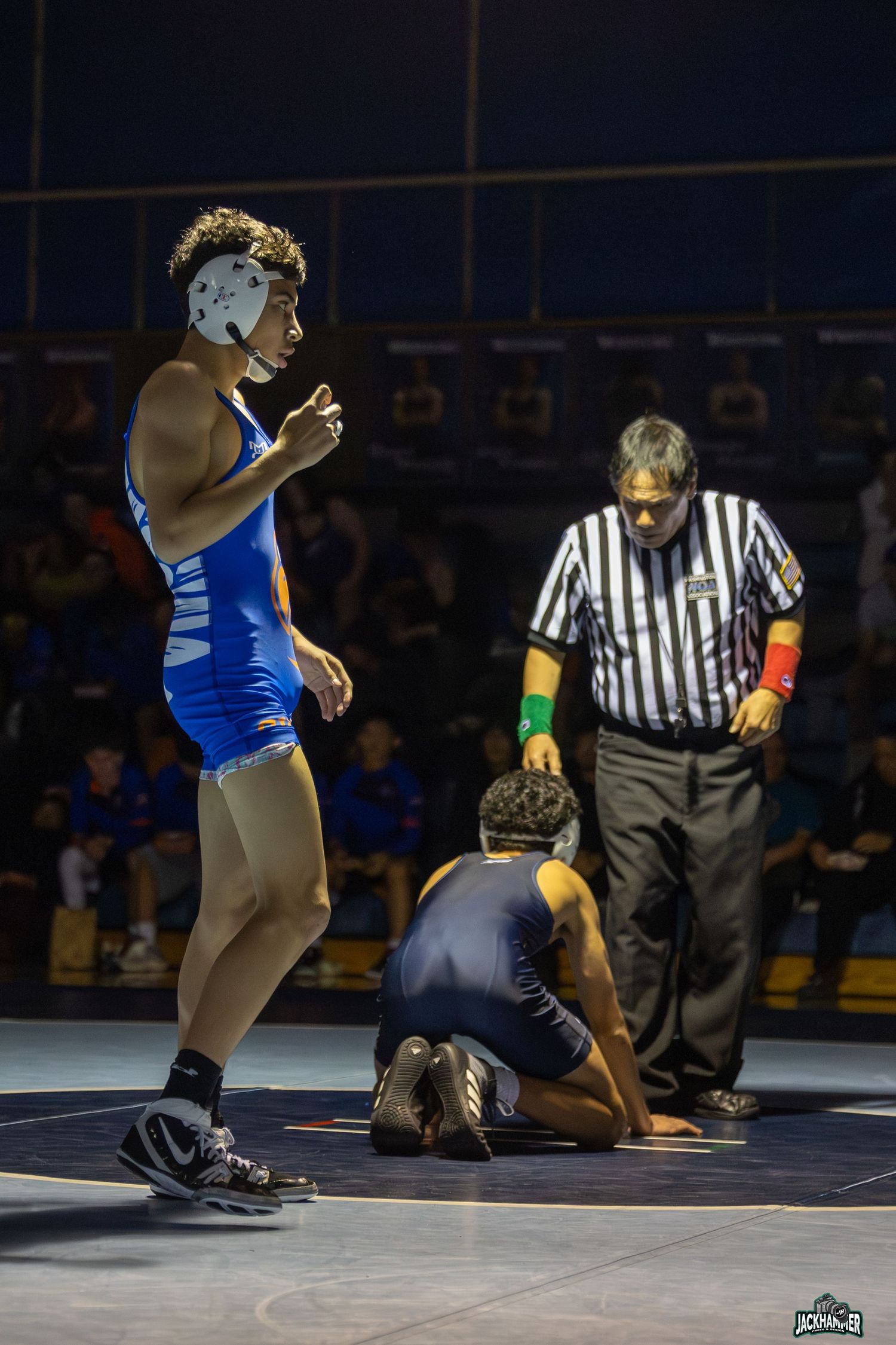 Wrestler in blue singlet stands victorious as opponent kneels on mat with referee nearby.