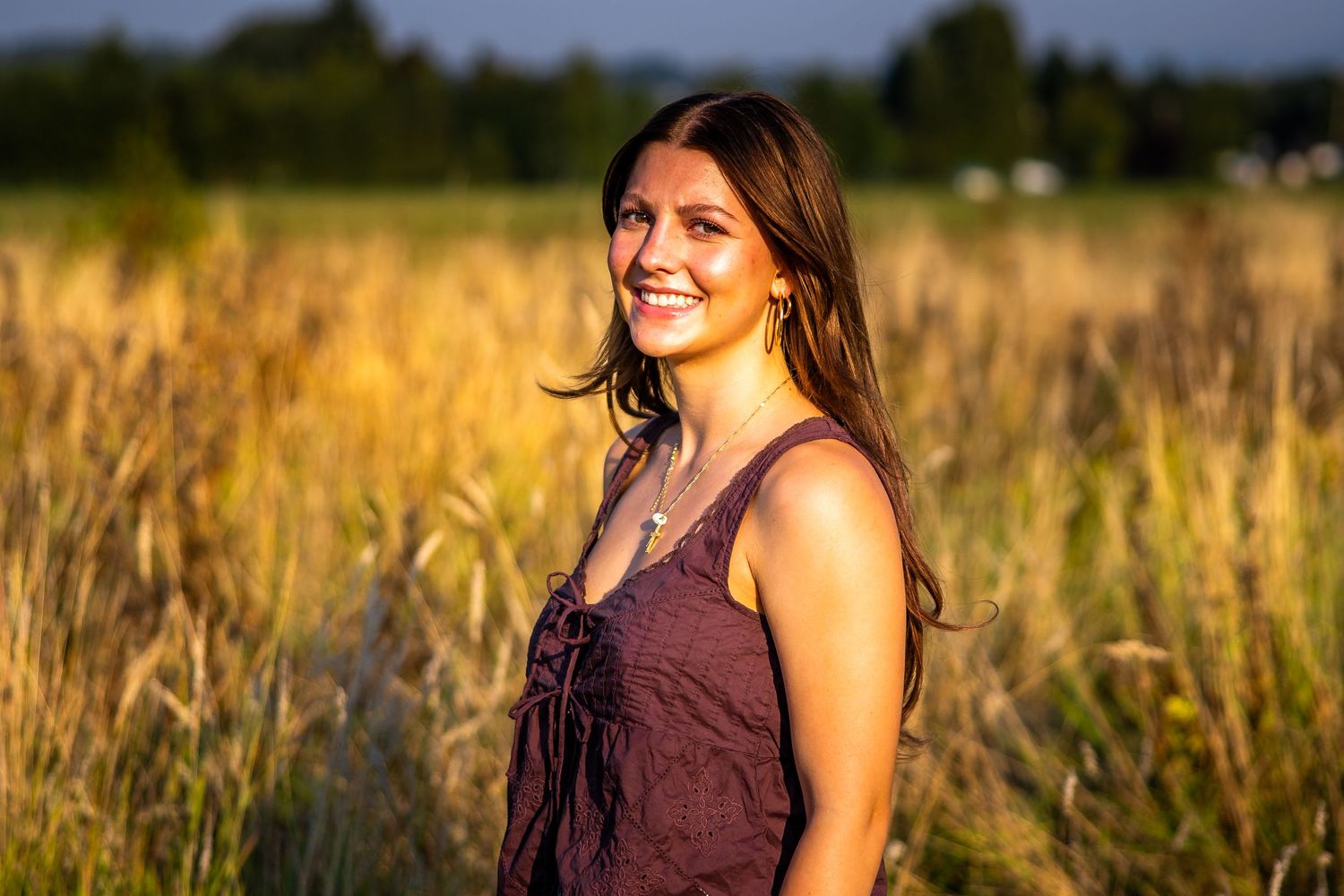 Smiling young woman in maroon dress standing in a golden sunlit field at sunset.