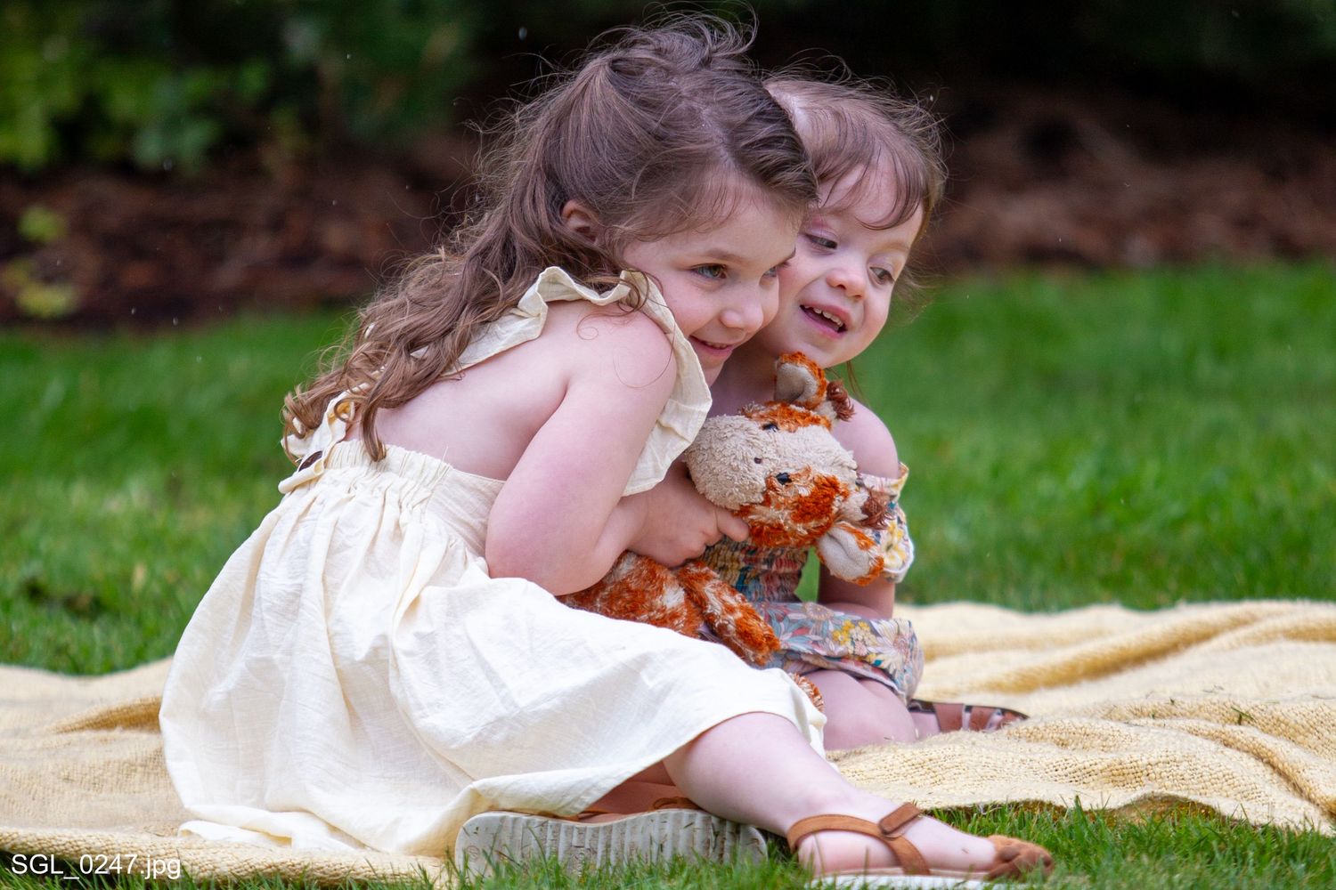 Two toddler girls in dresses sit on a blanket in the grass, hugging and playing outdoors.