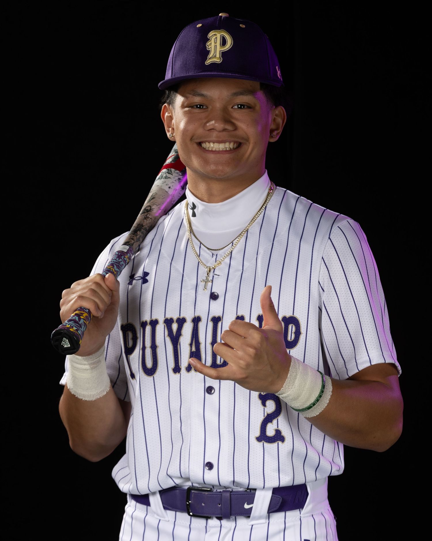 Puyallup baseball player #2 in purple pinstripe uniform smiles holding bat over shoulder giving thumbs up.