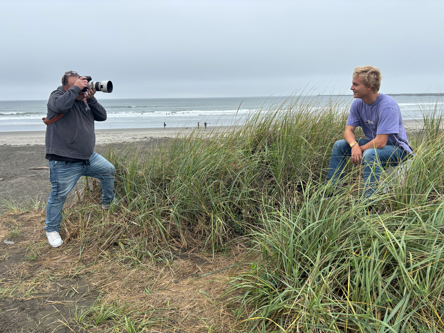 Photographer with telephoto lens shooting a young man sitting on beach grass dunes by the ocean.
