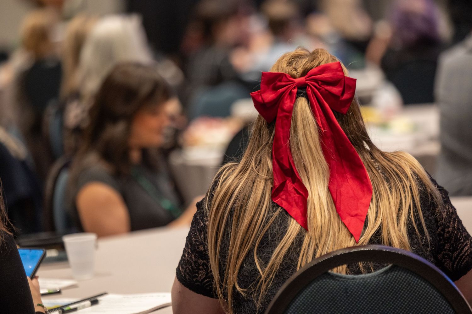 Back view of woman with long blonde hair wearing a large red bow at an indoor conference.