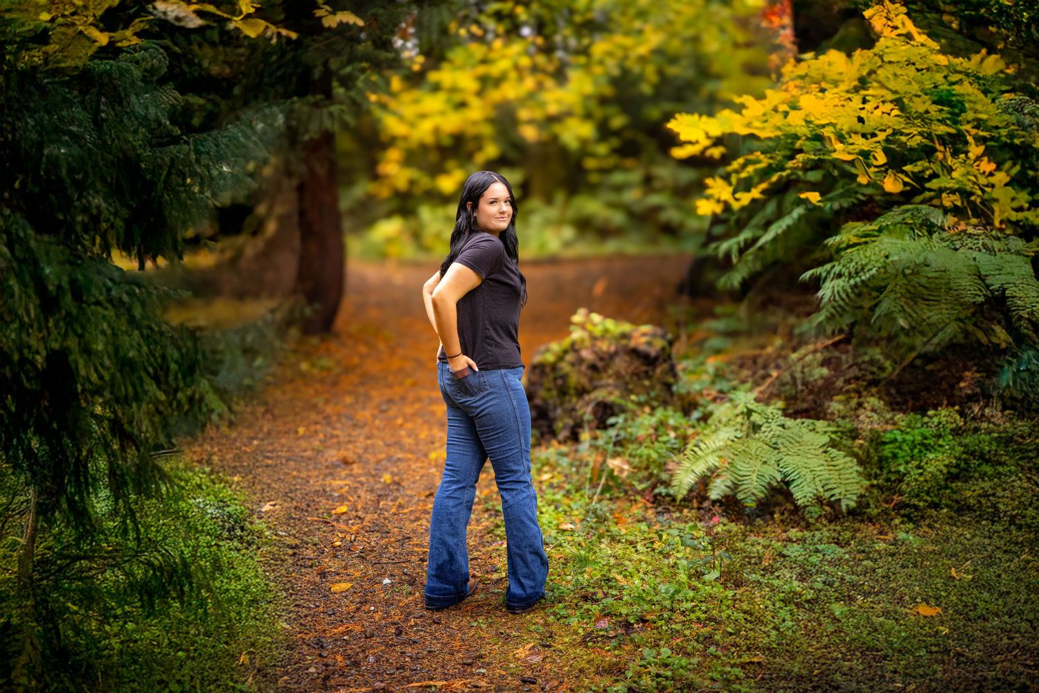 Teen girl in flare jeans posing on a forest path surrounded by autumn foliage.