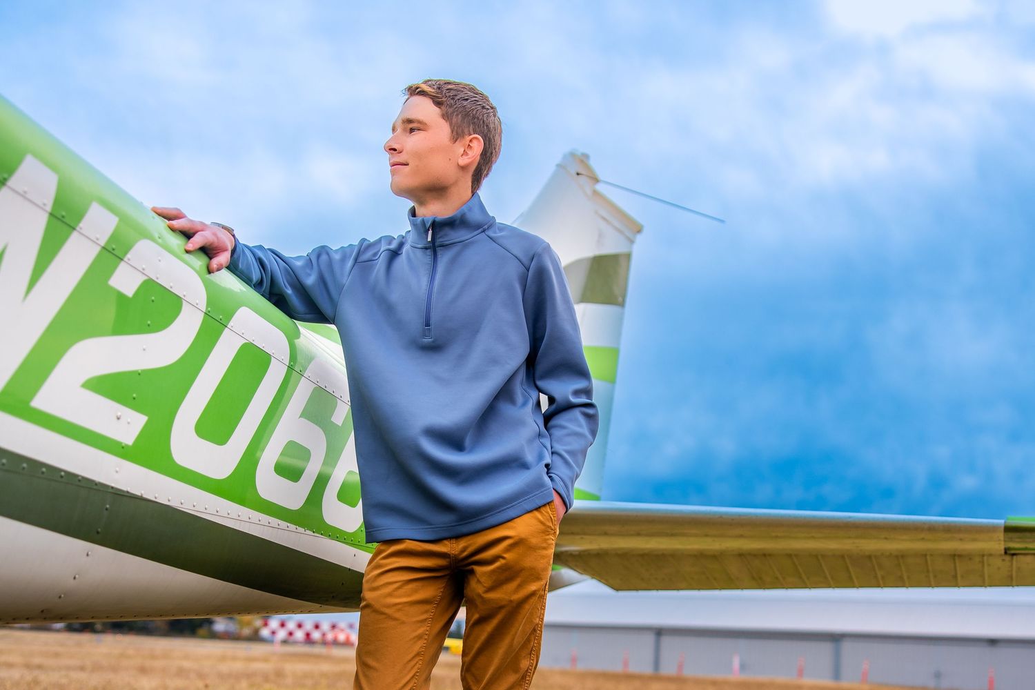 Young man in blue quarter-zip pullover standing beside a green airplane on a cloudy day.