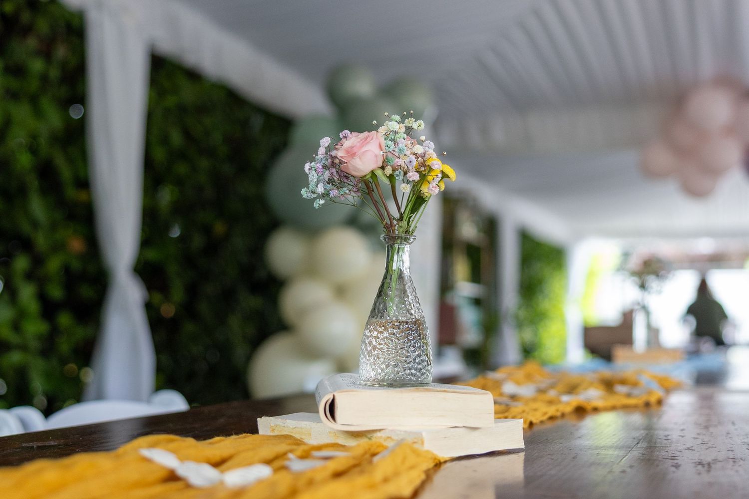 Glass vase with wildflowers on a book atop a wooden table at an outdoor event.