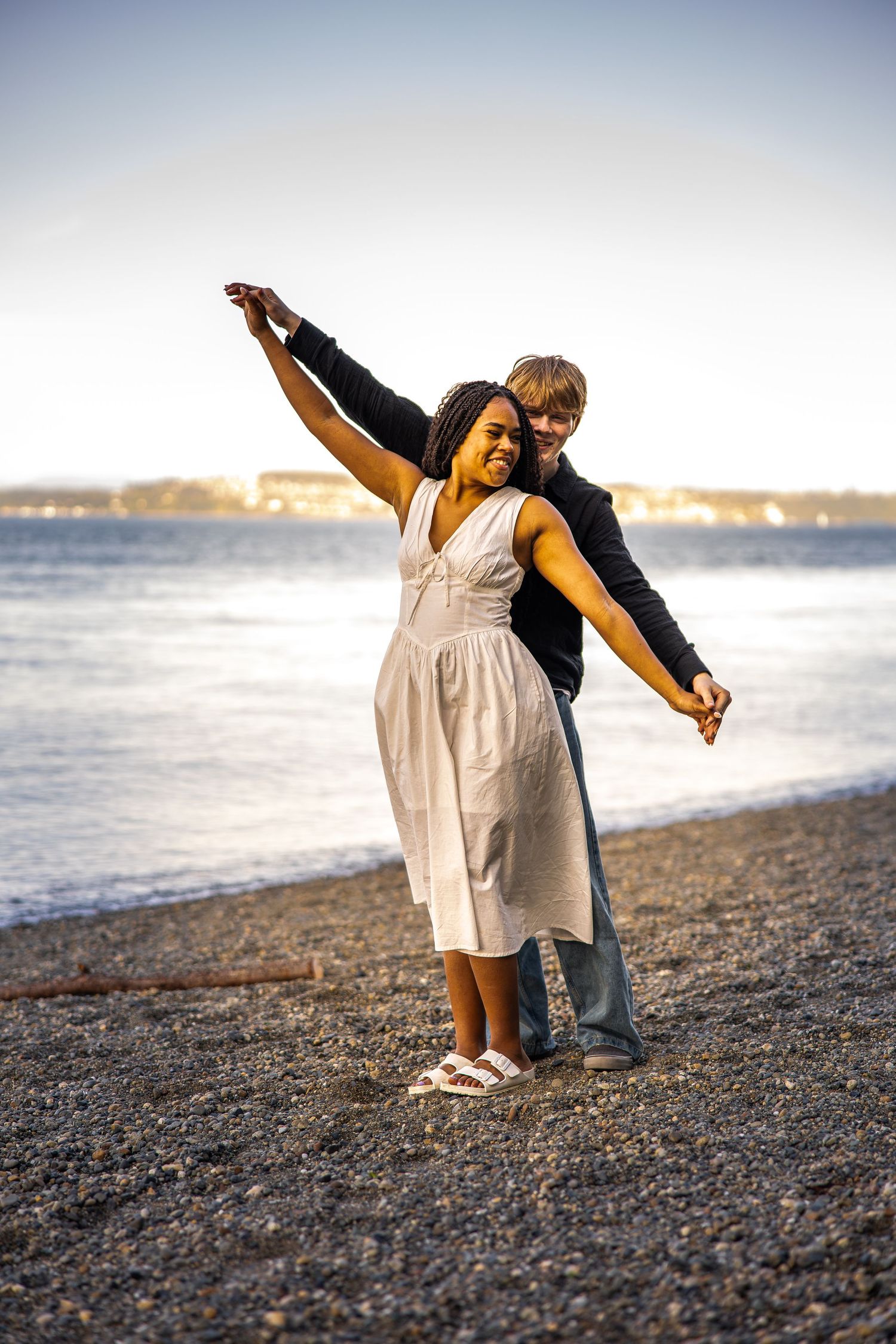 Young interracial couple dancing joyfully on a rocky beach at sunset near calm water.
