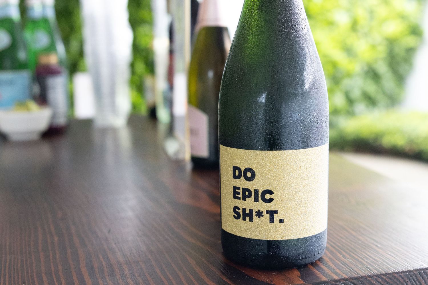 Wine bottle with bold label reading Do Epic Sh*t on wooden table at outdoor event.