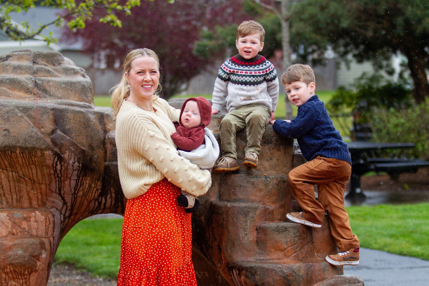 A smiling mother holds an infant while two young boys climb a decorative bear sculpture in a park.