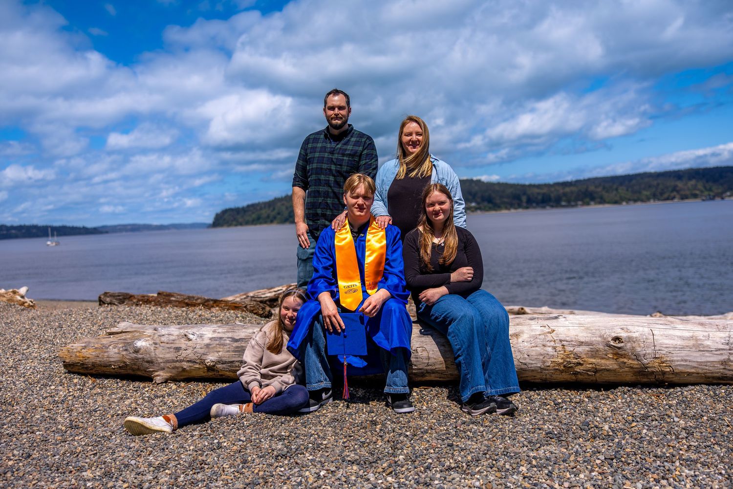 Family of five posing on driftwood log at rocky Pacific Northwest beach with blue sky and forested shoreline.