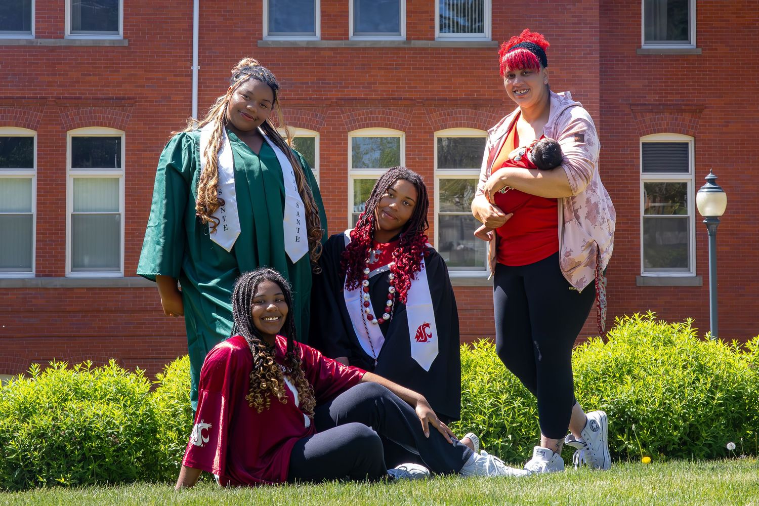 Four women celebrating graduation posing on green grass in front of a red brick university building.