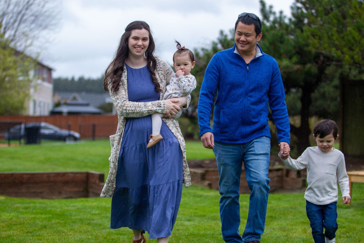 A happy family of four walks together on a green lawn, with a mother holding a baby and a toddler nearby.