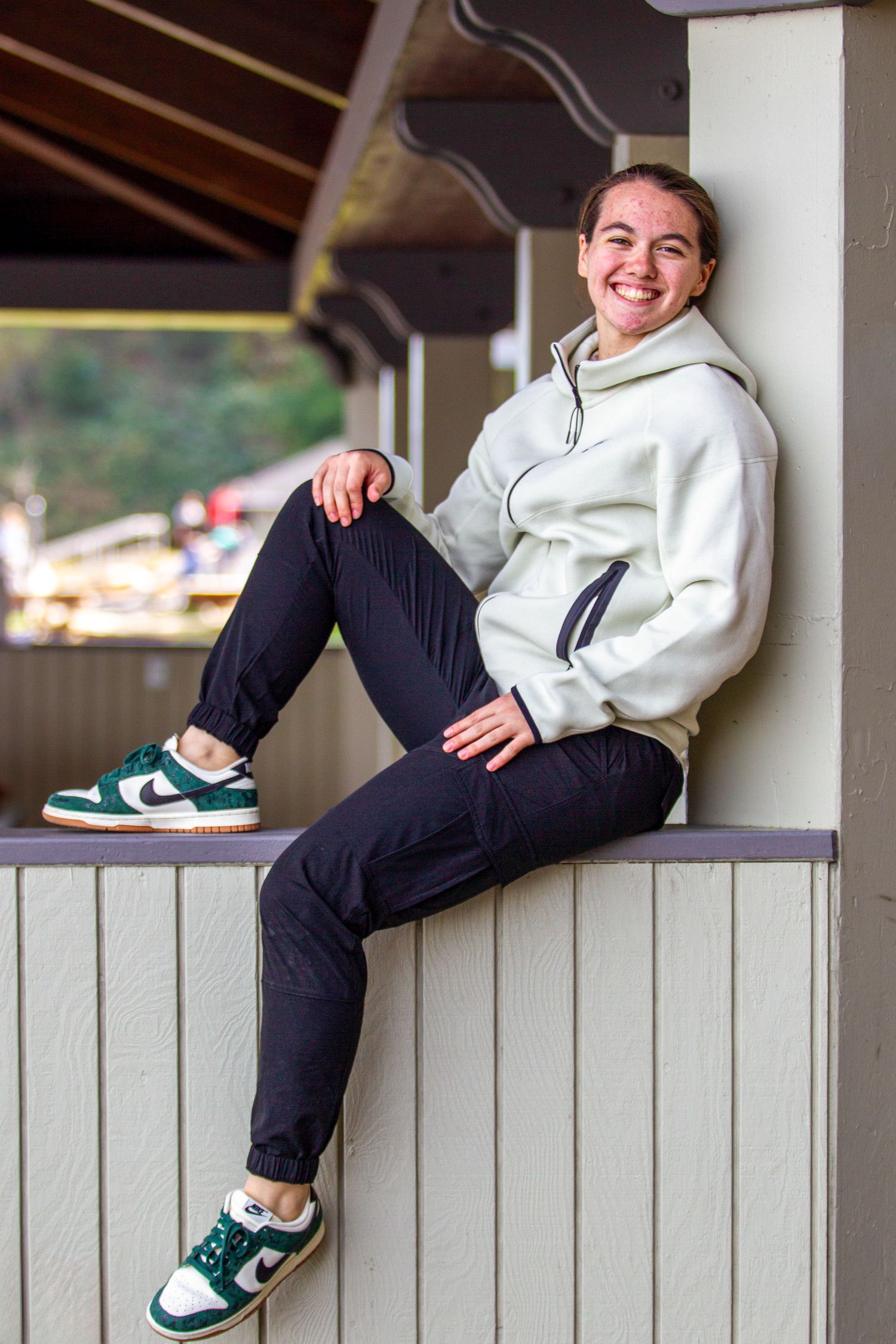 Smiling teen girl in white hoodie and Nike sneakers sitting on a ledge outdoors for senior photos.
