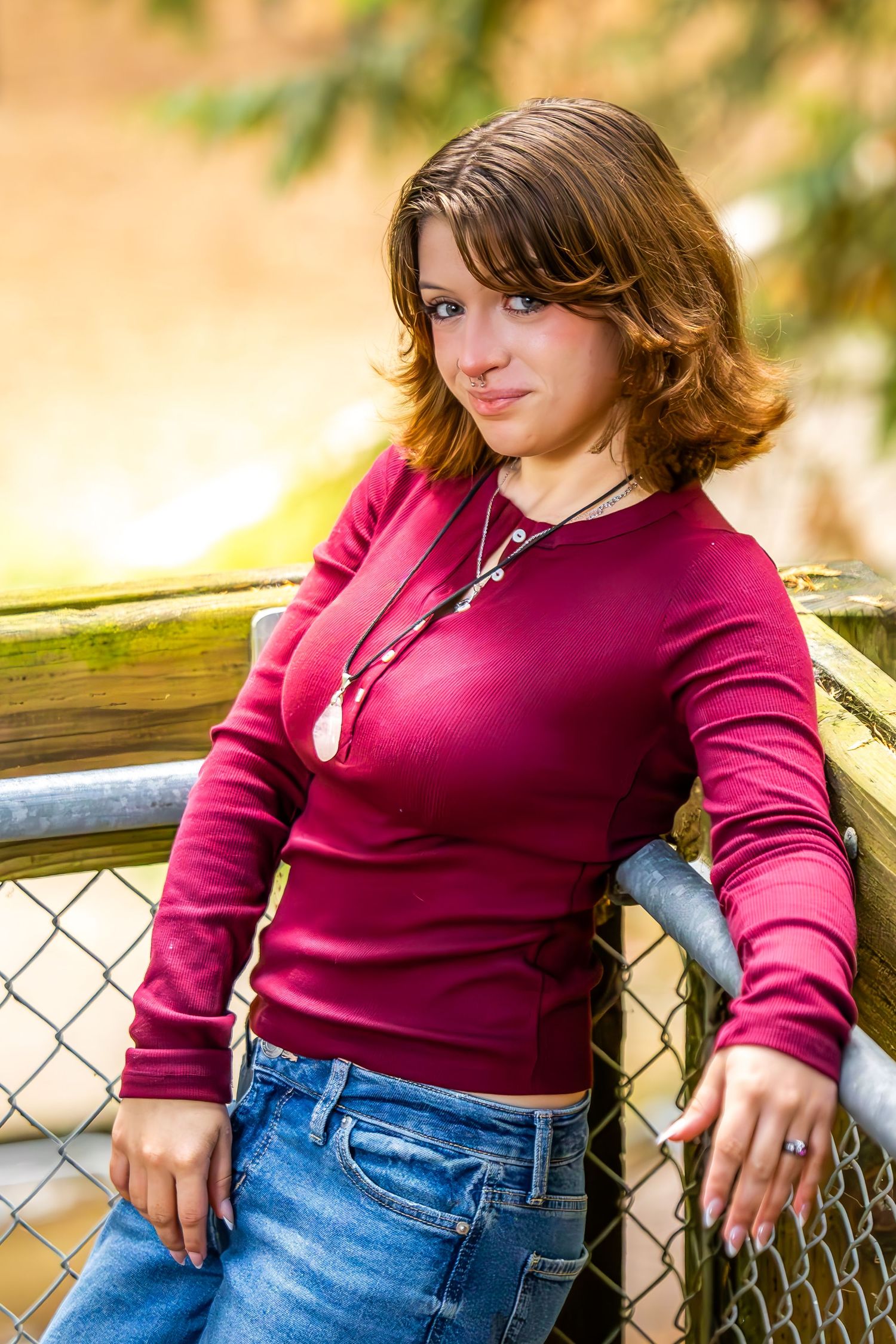 Young woman in burgundy top and jeans leaning against a chain-link fence in warm autumn light.
