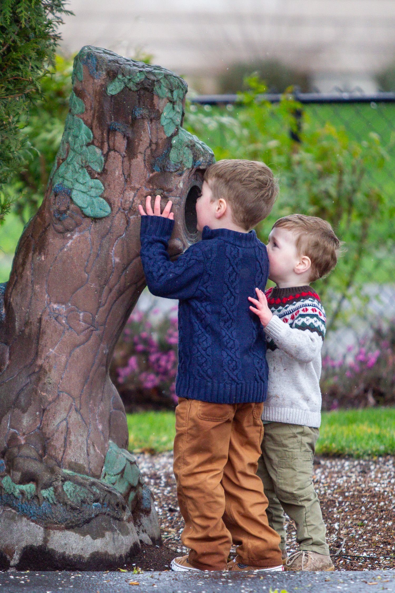 Two young boys in colorful sweaters explore a decorative tree sculpture in a garden setting.