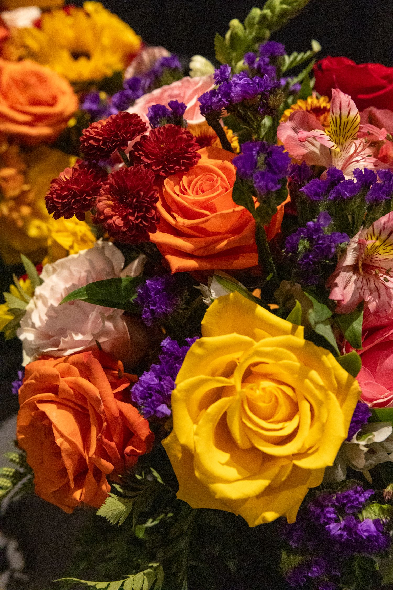 Vibrant bouquet of colorful roses including yellow, orange, and pink blooms with purple statice flowers.