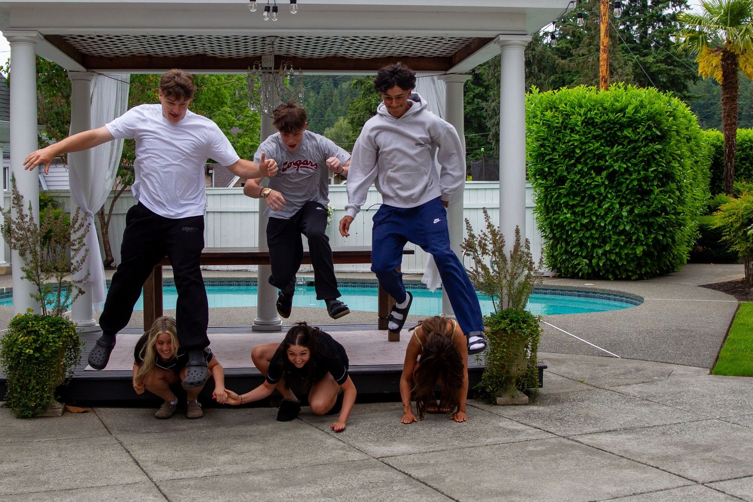 Group of young people jumping off outdoor steps near a swimming pool in a backyard.