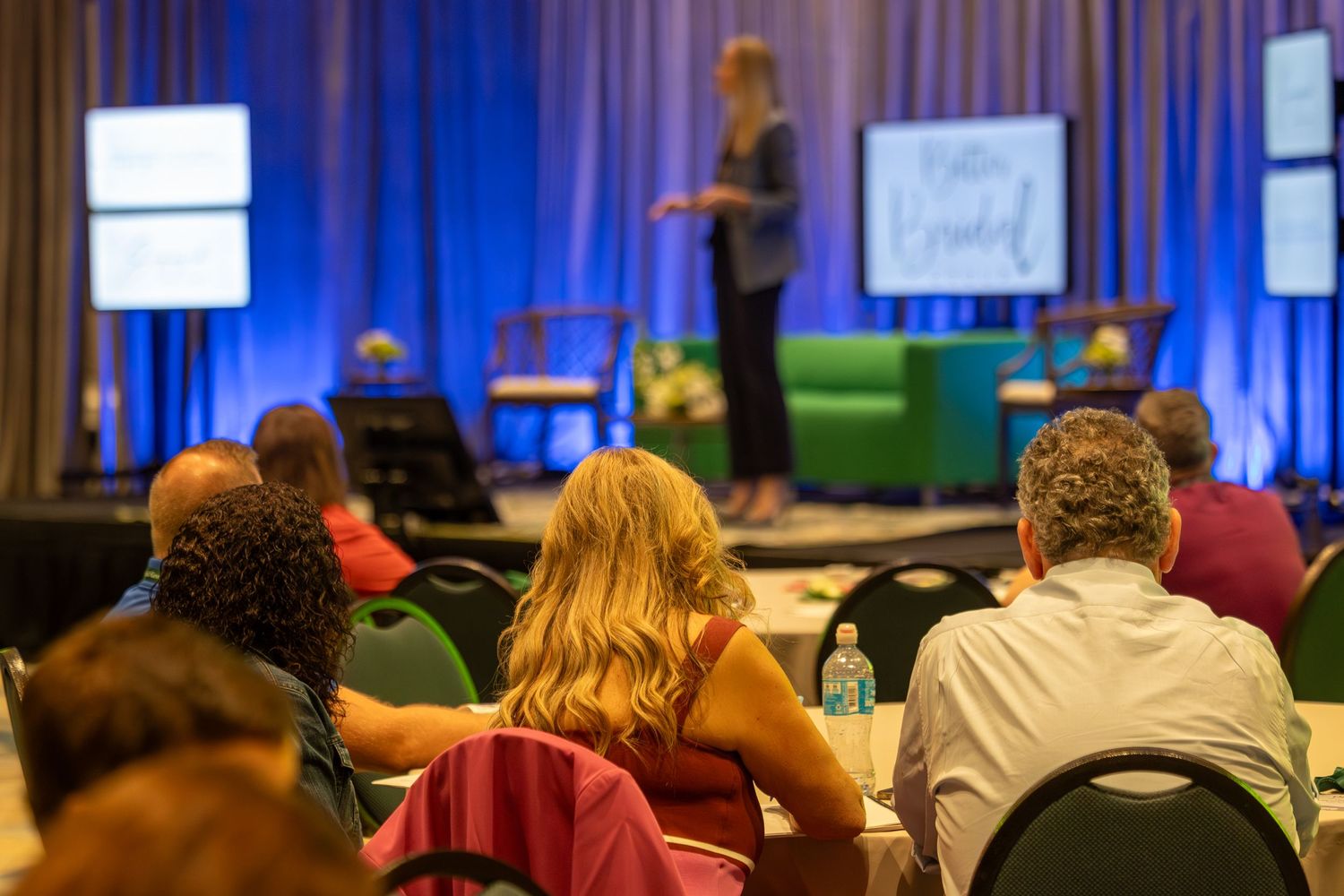 Audience members seated watching a female presenter on a blue-lit stage at an indoor conference event.