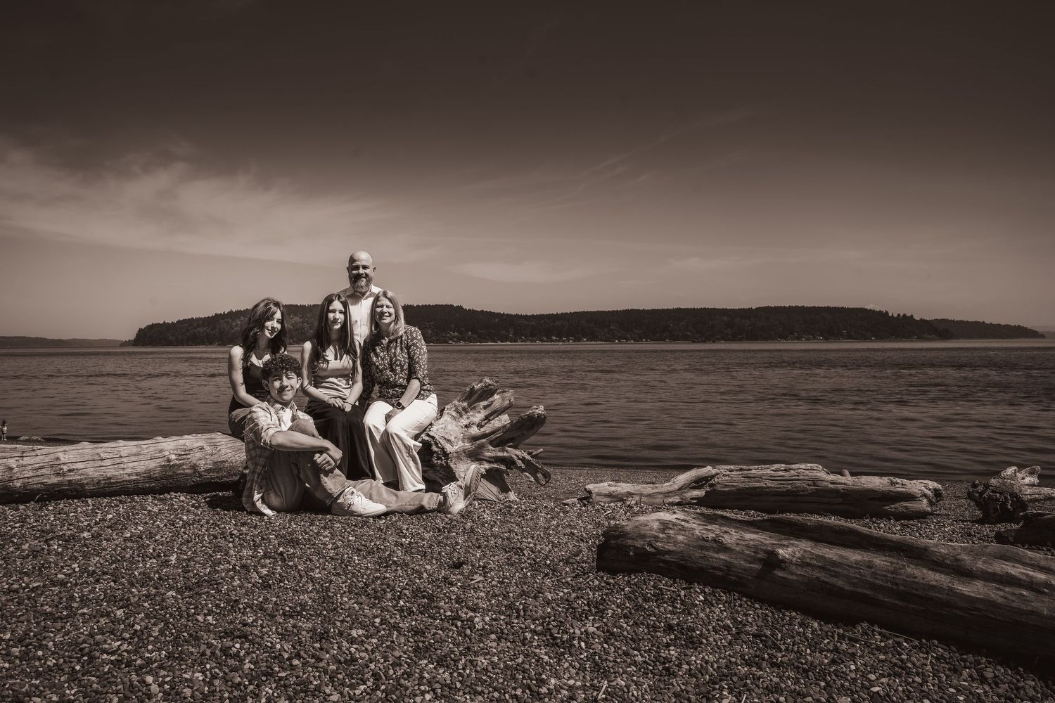 Sepia-toned family portrait on a rocky beach with driftwood logs and calm water with island in background.