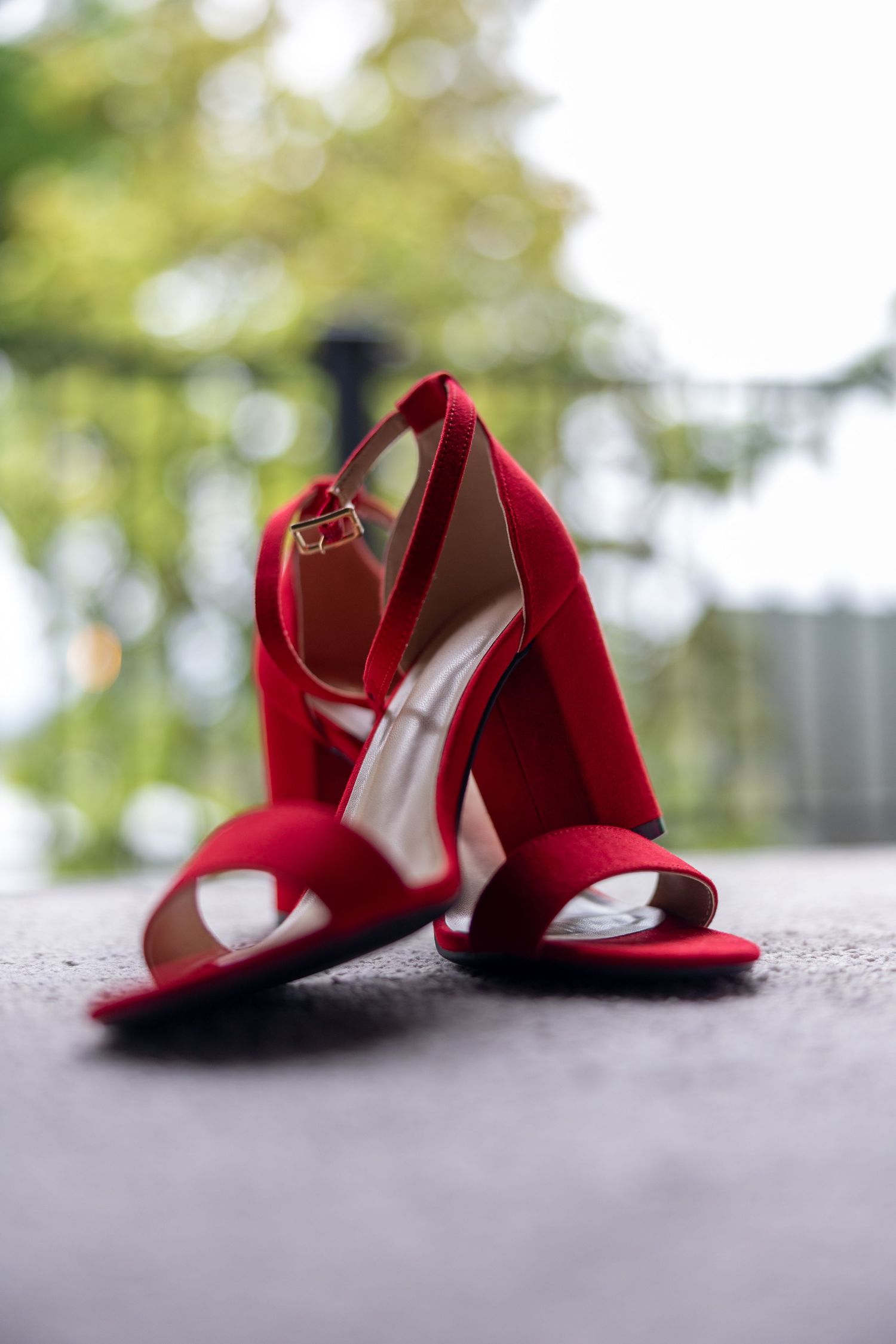 Red suede high heel sandals with ankle strap placed on concrete surface outdoors.